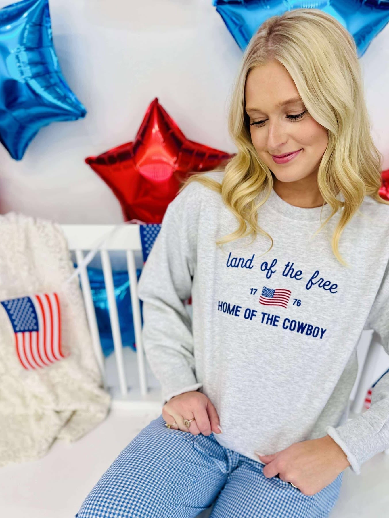 A woman sitting in front of a backdrop with American flag elements, wearing a gray sweatshirt with the text 'Land of the Free Home of the Cowboy' and stars.