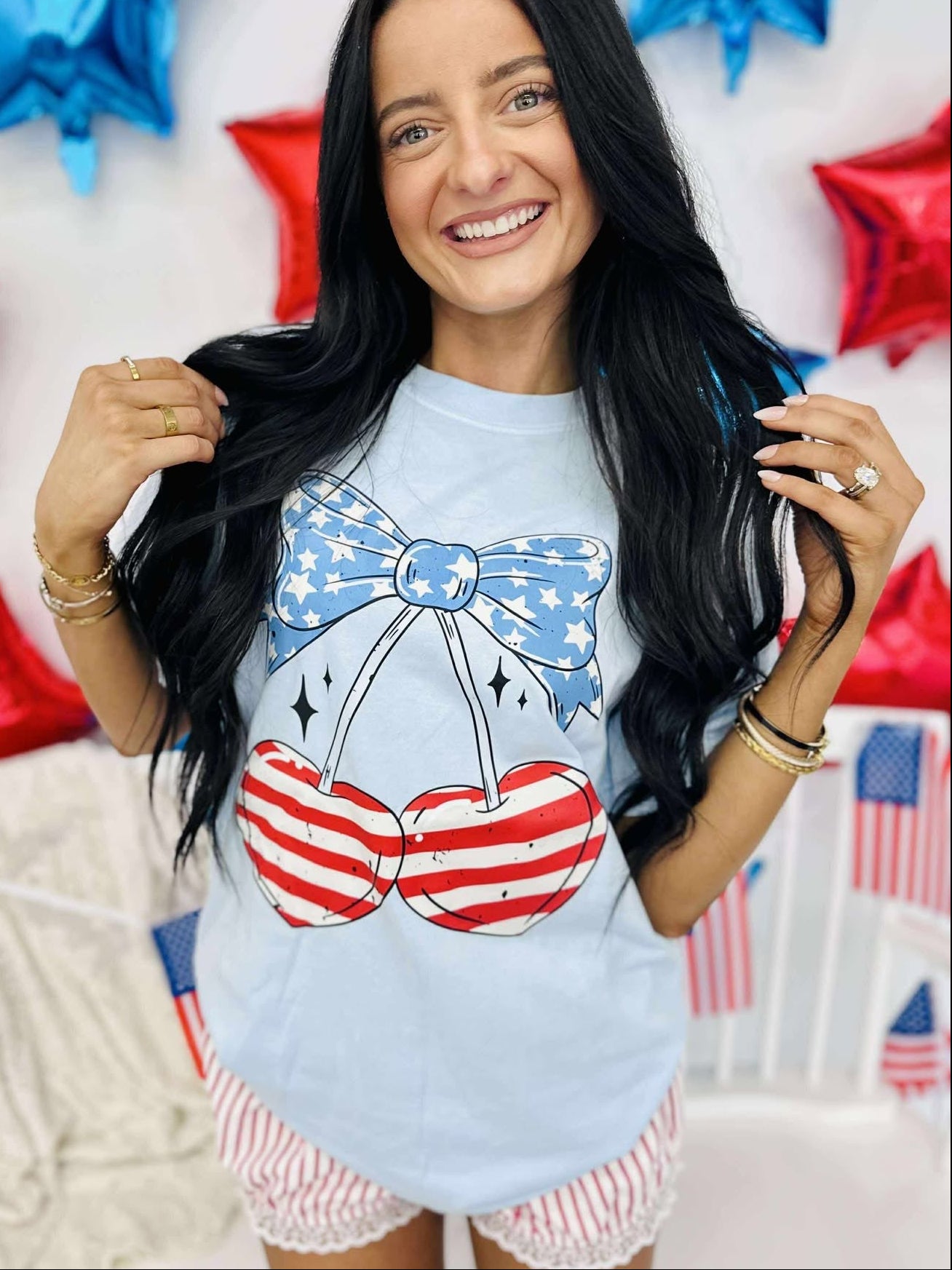 A person wearing a light blue graphic tee with a patriotic cherry design, shorts, and standing in front of a backdrop with red and blue stars.