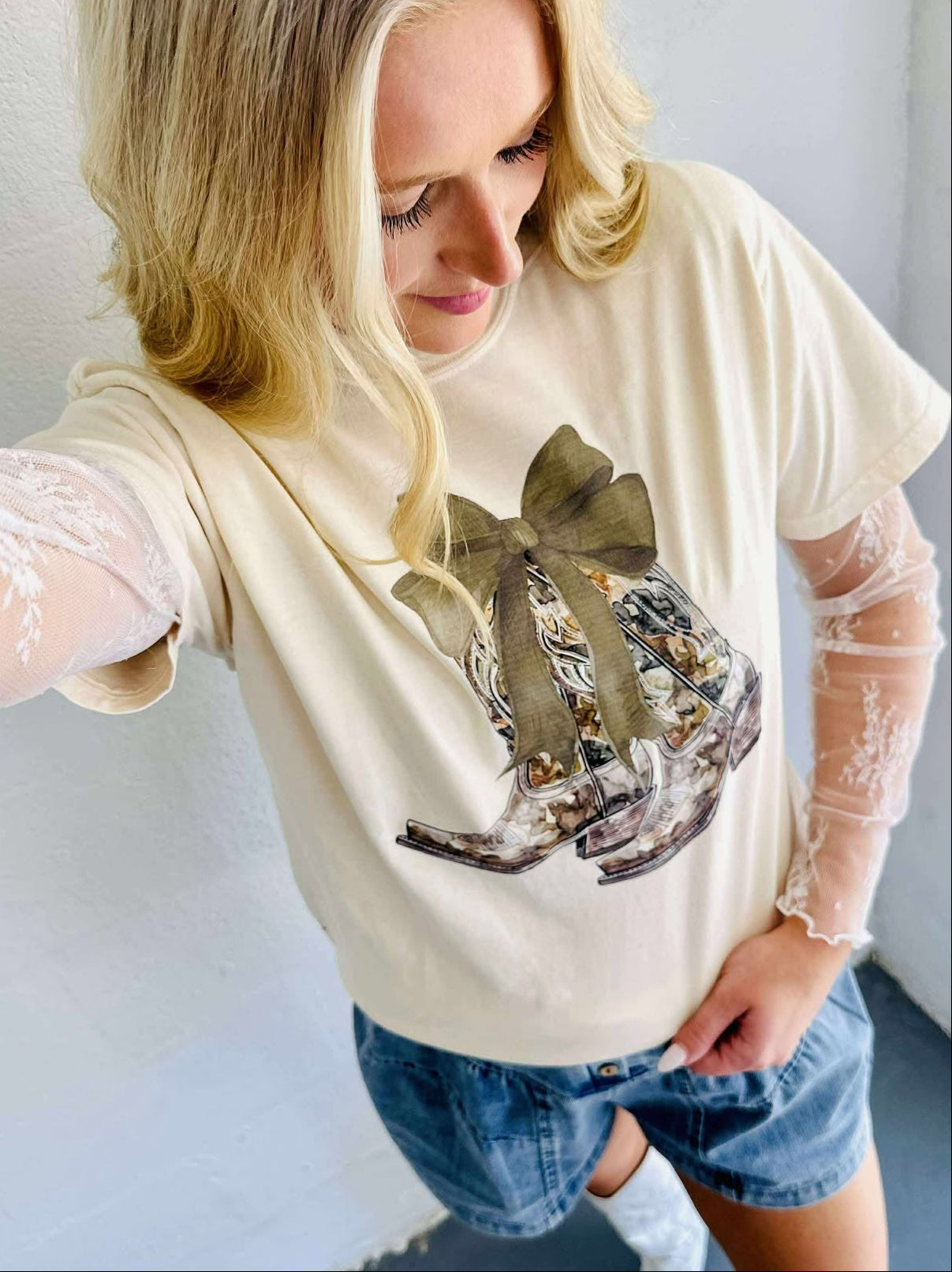 A graphic tee with a camo bow and boots design, predominantly in beige color, paired with blue denim shorts and white boots.