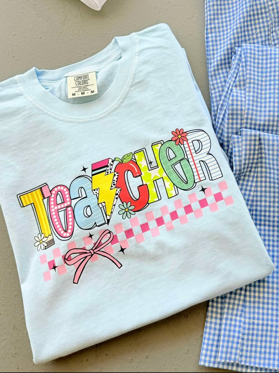 Folded t-shirt with 'TEACHER' text on a checkered fabric background