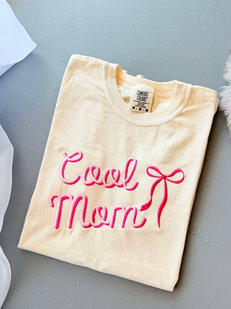 An ivory colored graphic tee with 'Cool Mom' embroidered in pink lettering, accompanied by a pink bow illustration.