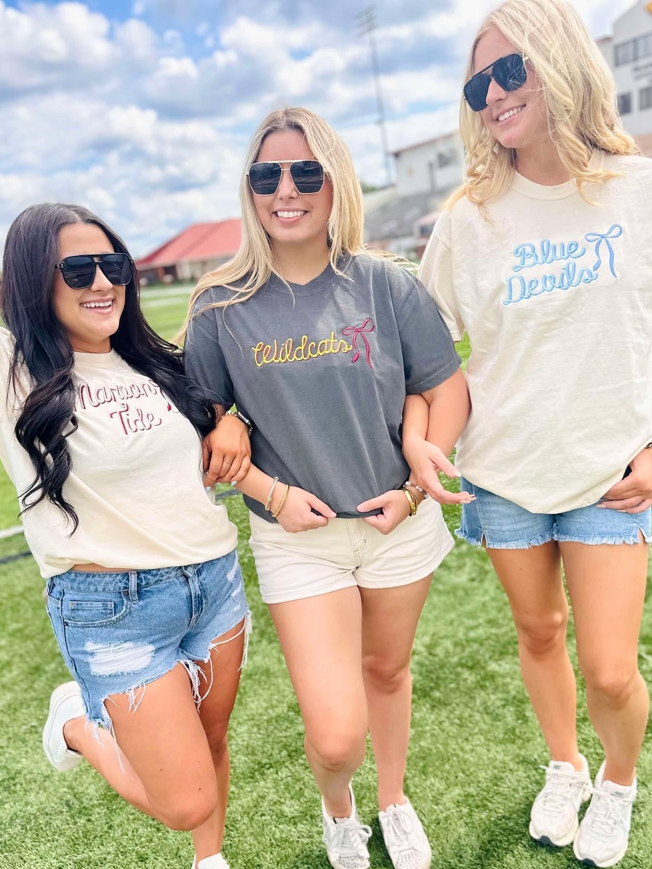 Three women standing on a grassy field wearing casual outfits with visible mascot school text on their shirts.