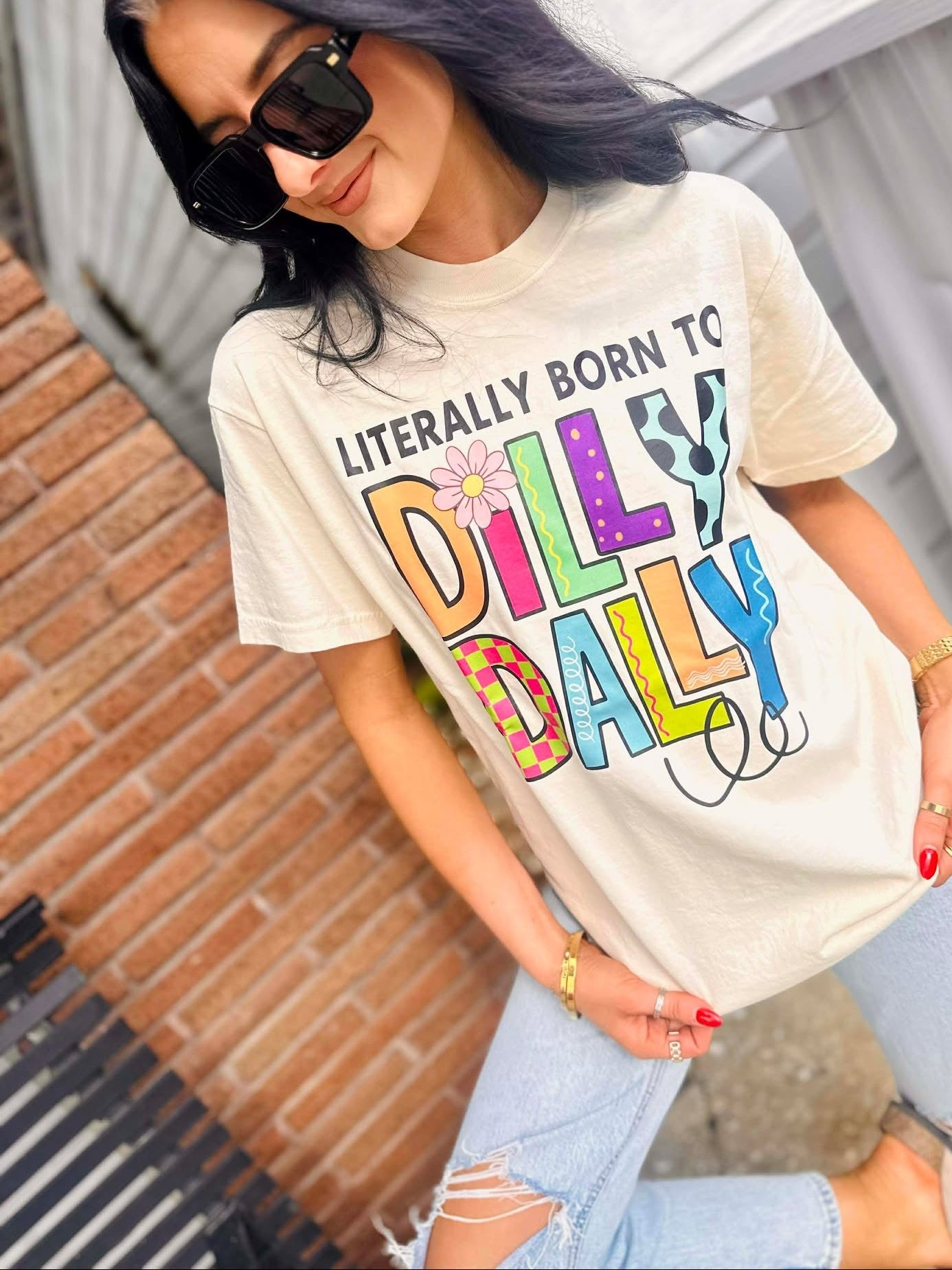 Woman wearing a Born to Dilly Dally Ivory t-shirt with colorful text outdoors
