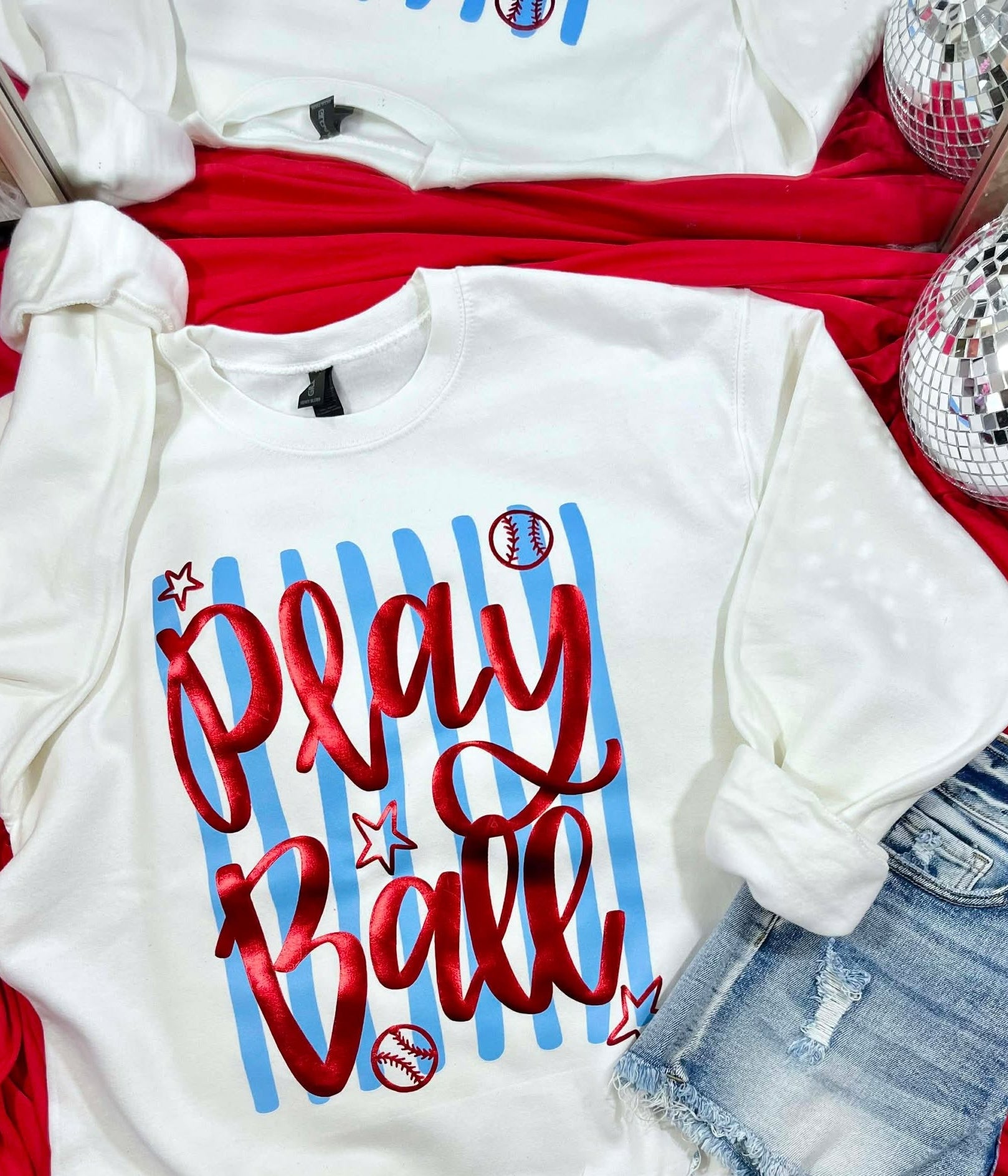 White sweatshirt with 'Play Ball' text on a red surface with denim shorts and disco balls.