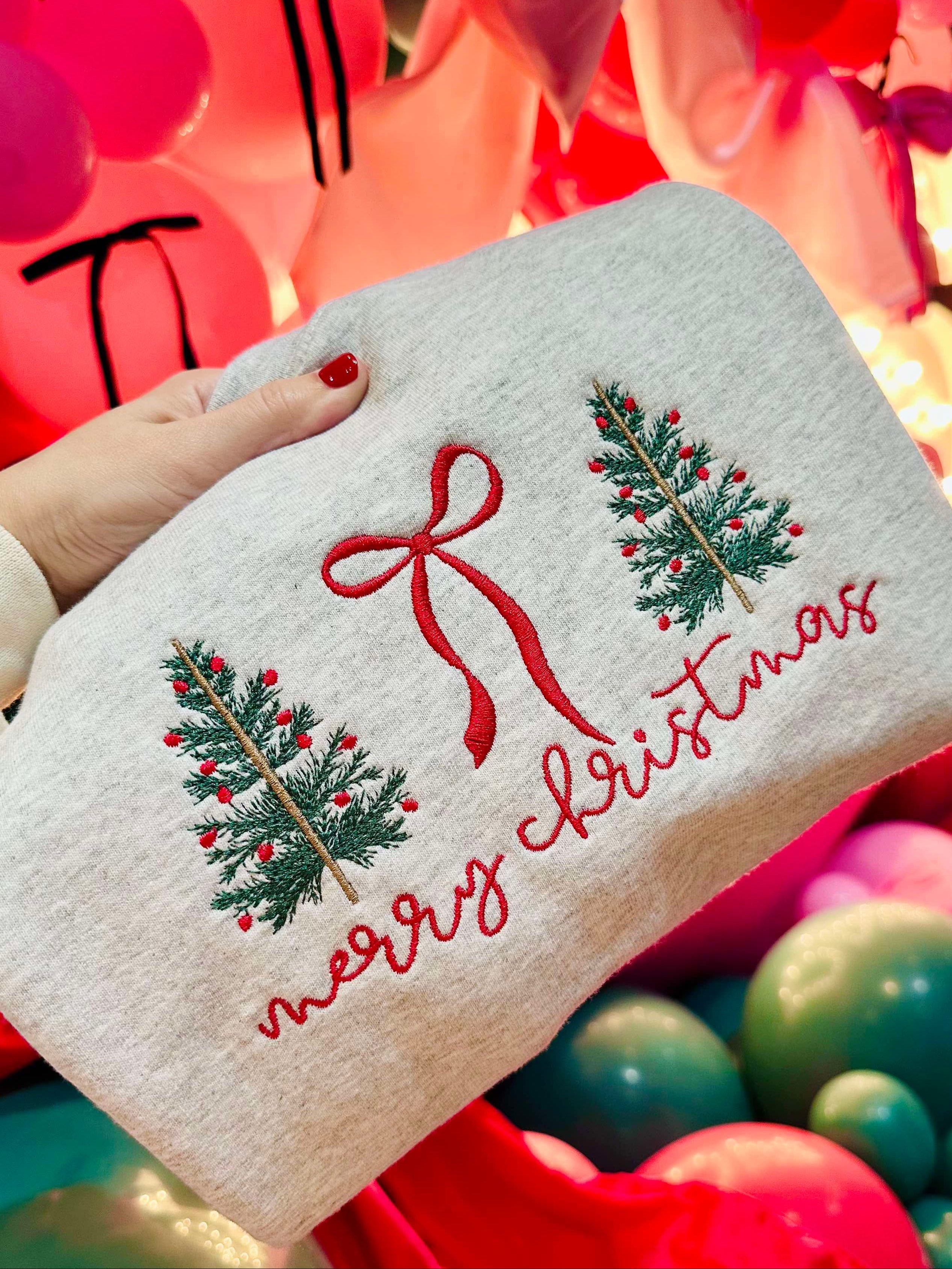 White sweatshirt folded with Christmas tree and bow design held by a hand against a colorful background of balloons.