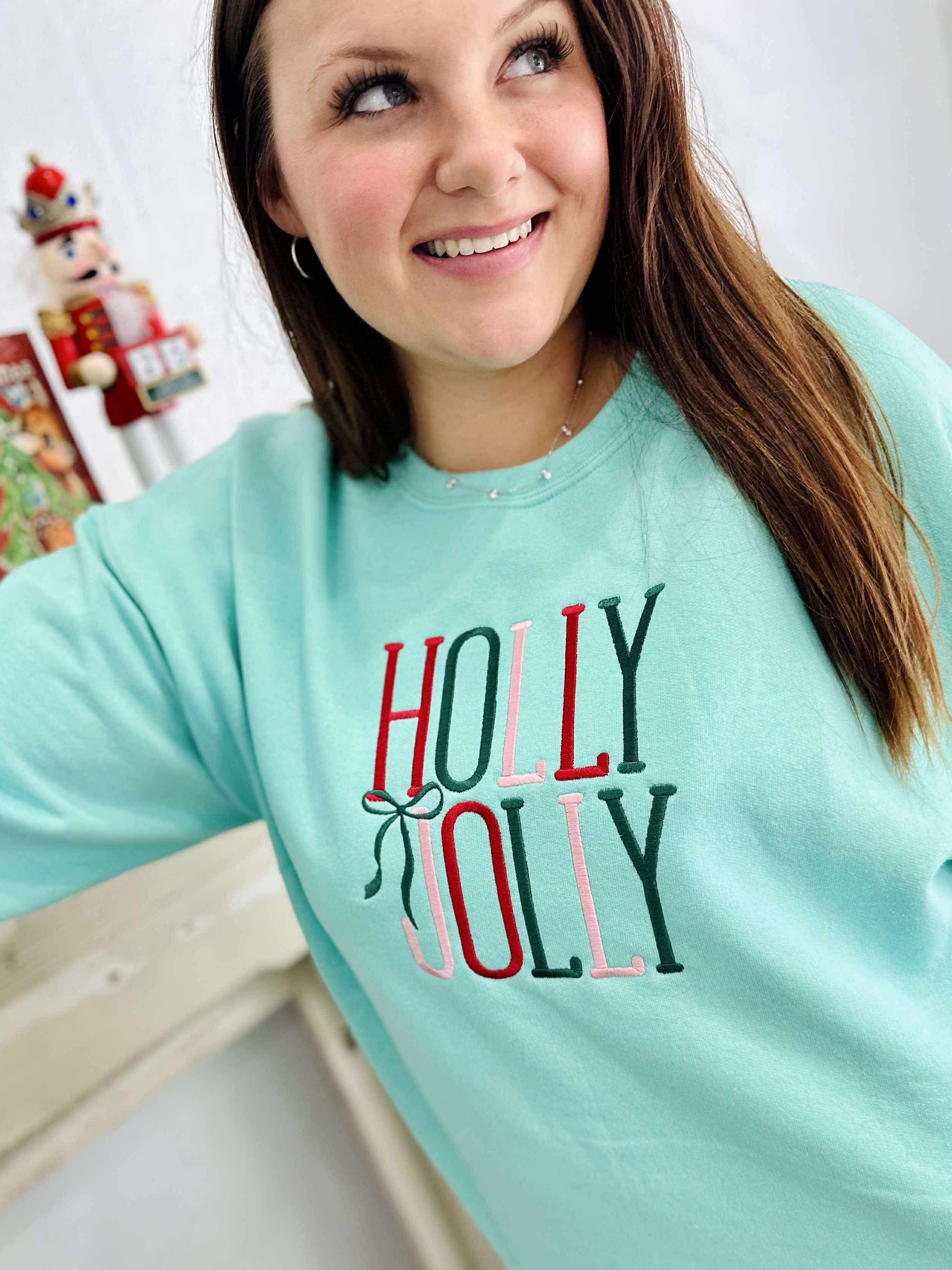 Woman wearing a light blue sweatshirt with 'Holly Jolly' thread, standing indoors.