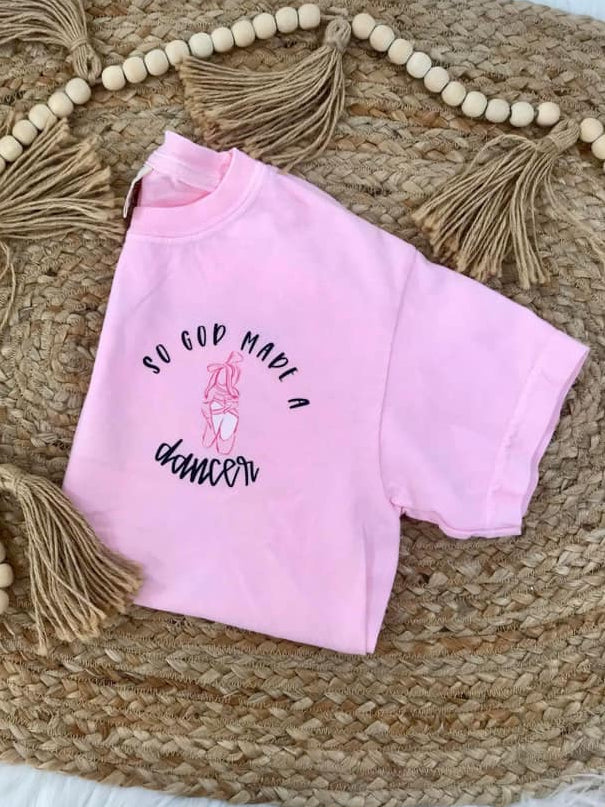 Pink t-shirt with 'So God Made a Dancer' text on a woven mat with beads and tassels.