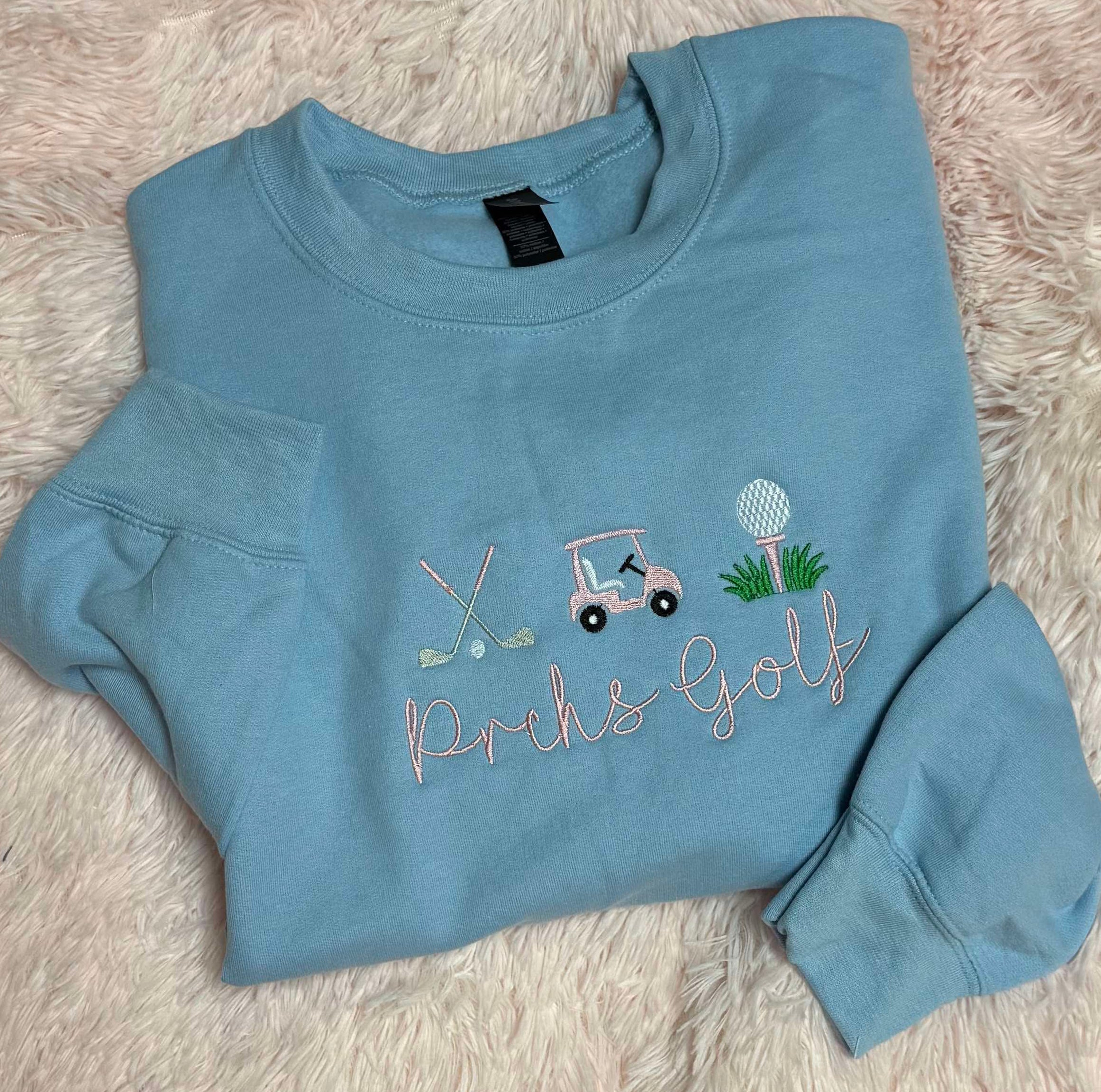 Blue sweatshirt with golf-themed school or custom design on a fluffy pink surface