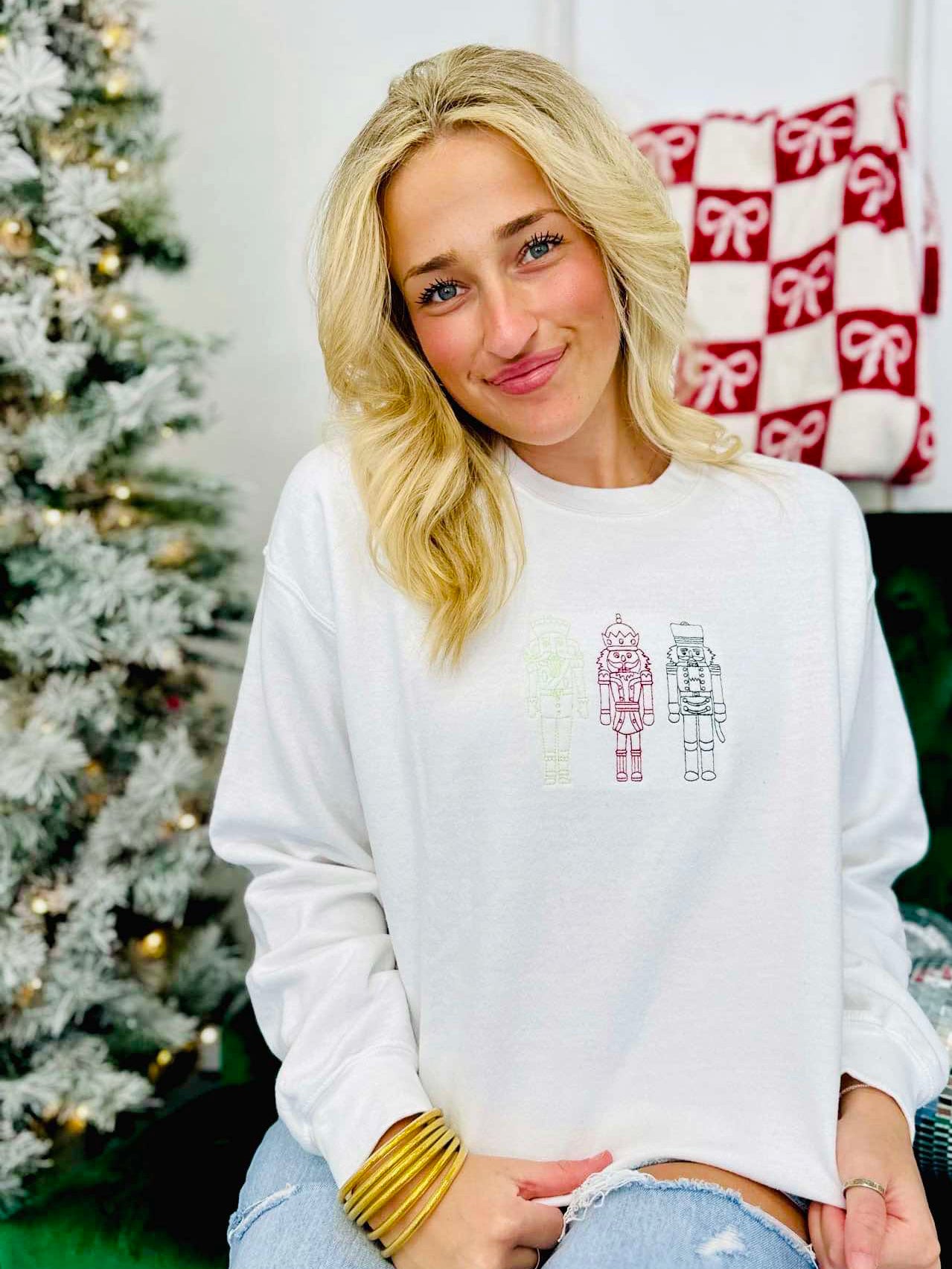 A woman sitting and wearing a white embroidered sweatshirt with the text 'Christmas Nutcracker Trio' on it, in front of a blurred Christmas tree and gifts in the background.