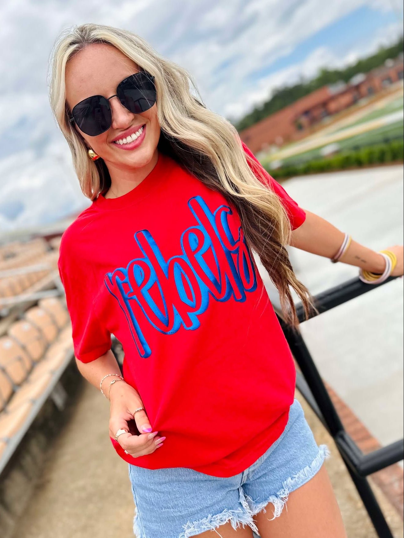 Woman wearing a red 'Rebels' shirt with sunglasses and denim shorts outdoors.