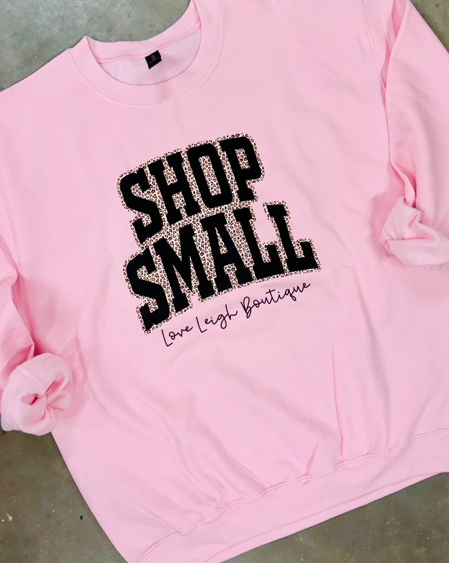 Pink sweatshirt with Leopard 'Shop Small' text on a concrete surface