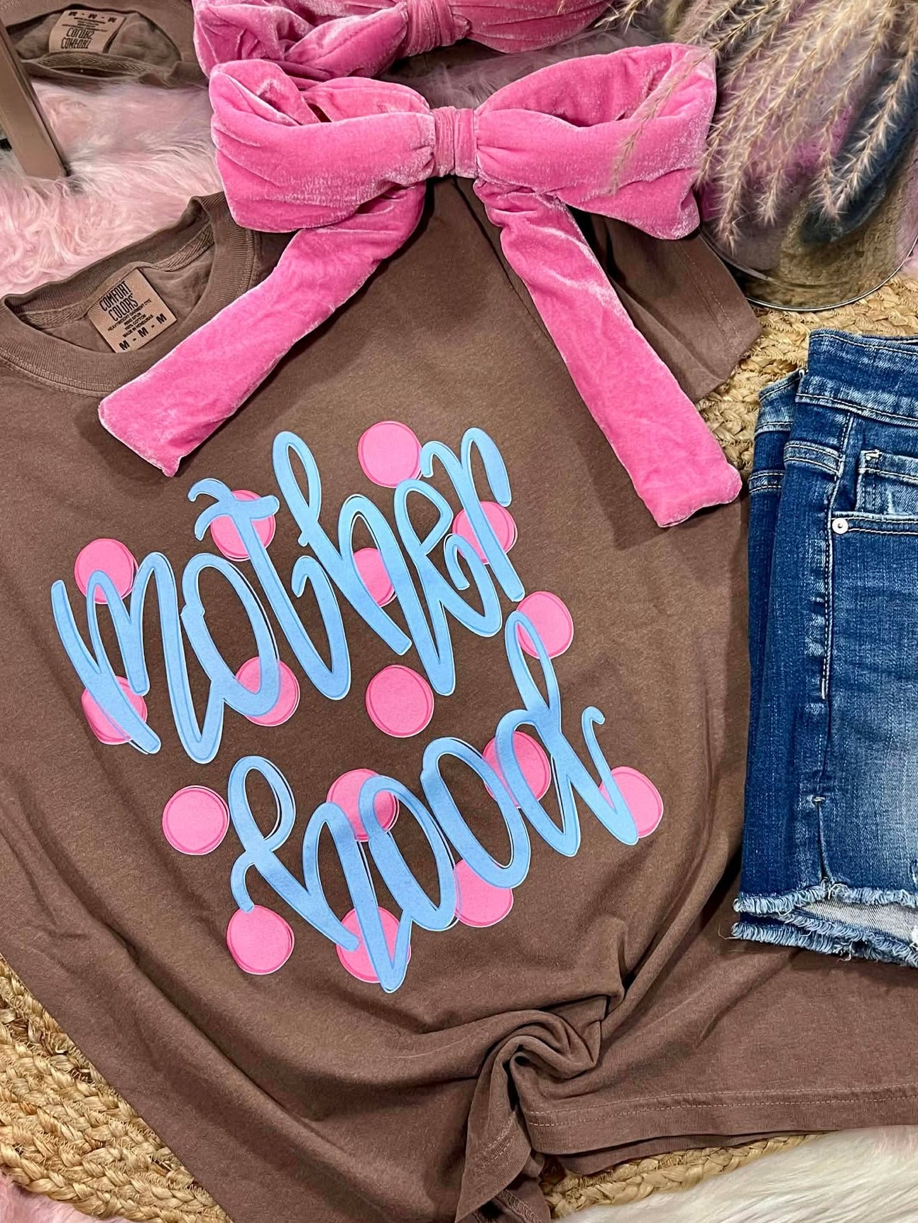 Brown t-shirt with 'Mother Hood' text and pink polka dots, paired with denim shorts and a pink bow on a textured surface.