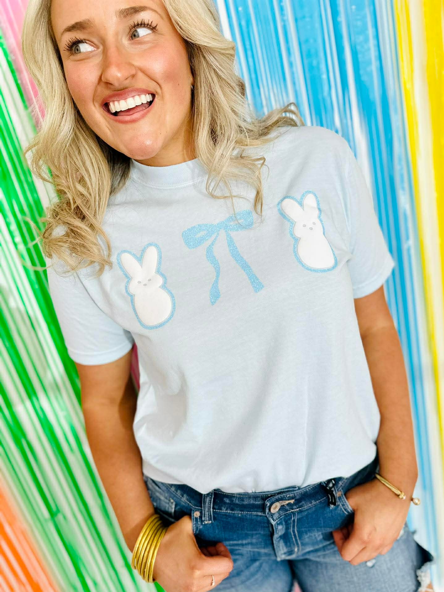 Person wearing a white t-shirt with blue designs with Puff Peeps Bunny and glitter bows against a colorful striped background