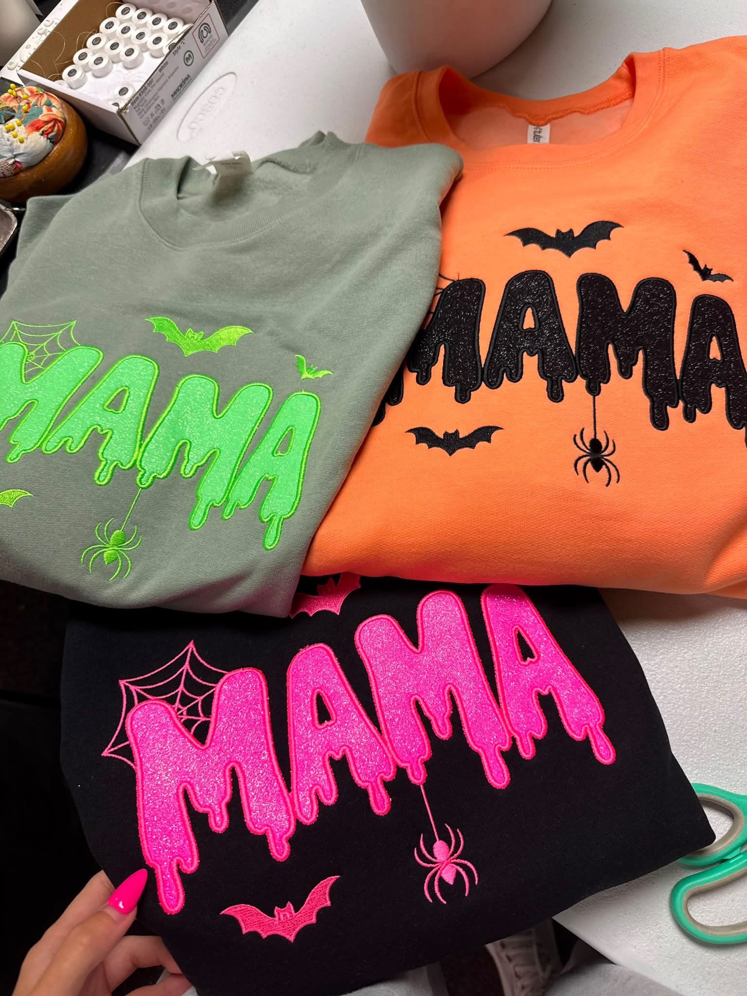 Three folded Halloween-themed t-shirts displayed together on a white surface. The top-left shirt is sage green with neon green dripping 'MAMA' text and small green spider/bat graphics.