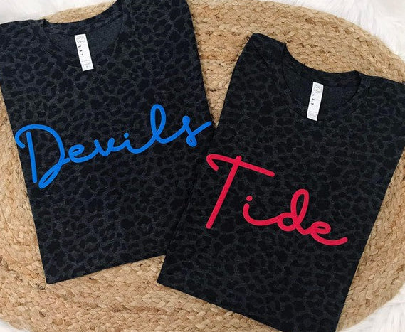 Two black leopard patterned t-shirts with 'Devils' in blue and 'Tide' in red on a woven surface.
