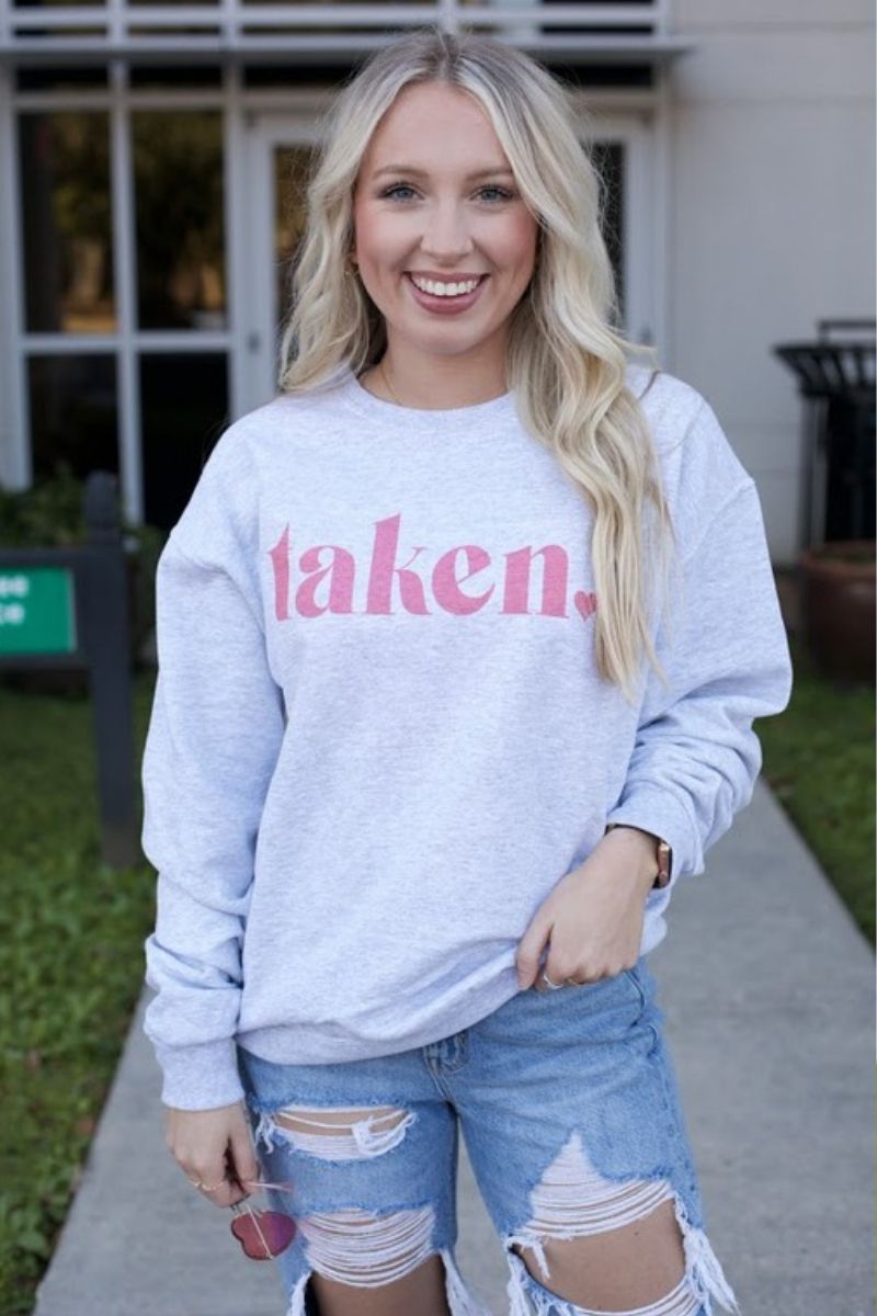 A woman standing outside wearing a gray sweatshirt with the text 'taken' printed in pink, paired with blue denim shorts with a frayed hem.