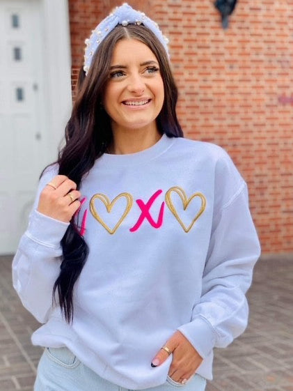 Woman wearing a white sweatshirt with heart and X puff glitter, standing in front of a brick wall.