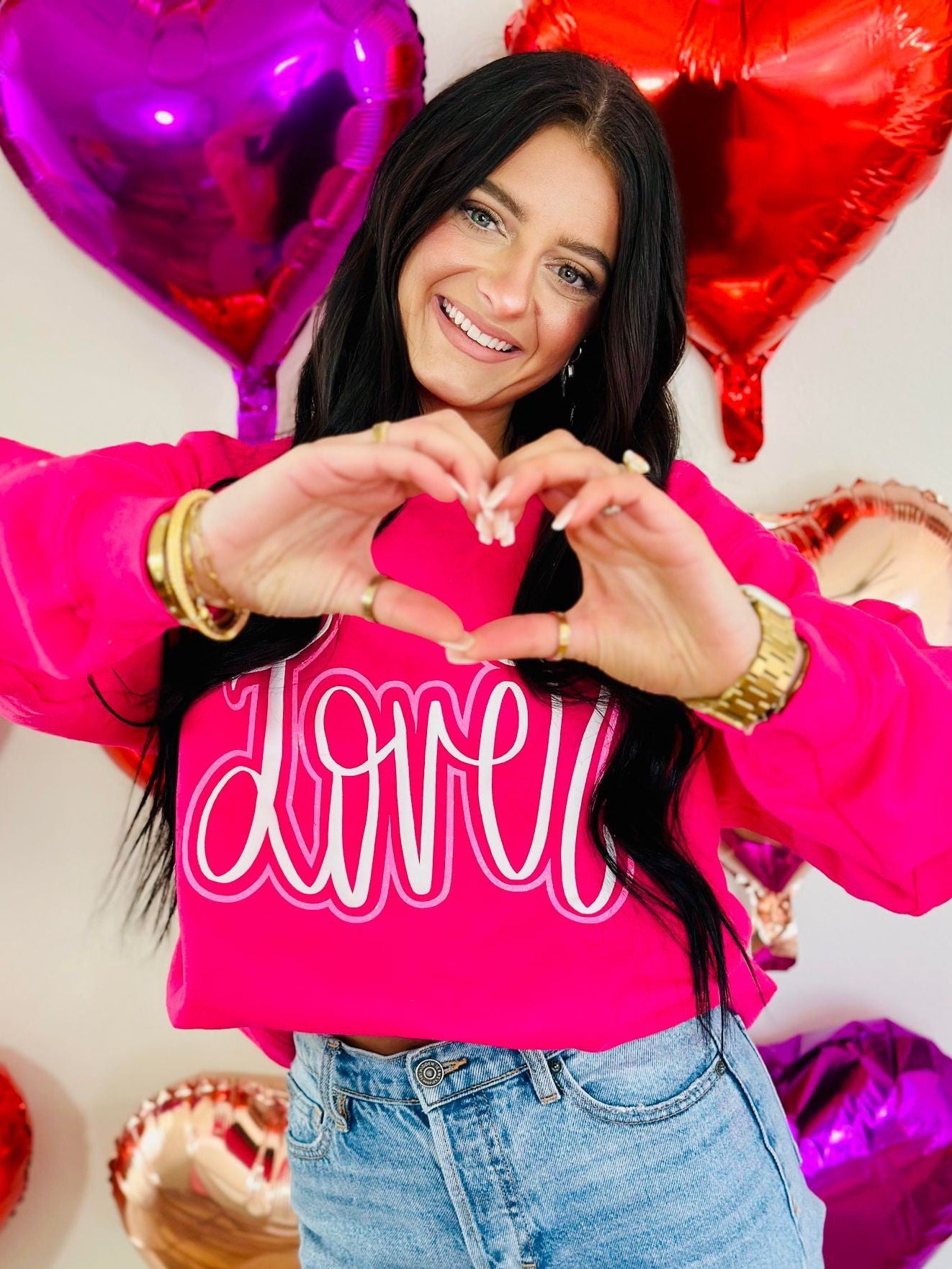 Woman in a pink sweatshirt with 'LOVER' text, making a heart shape with her hands, surrounded by heart-shaped balloons.