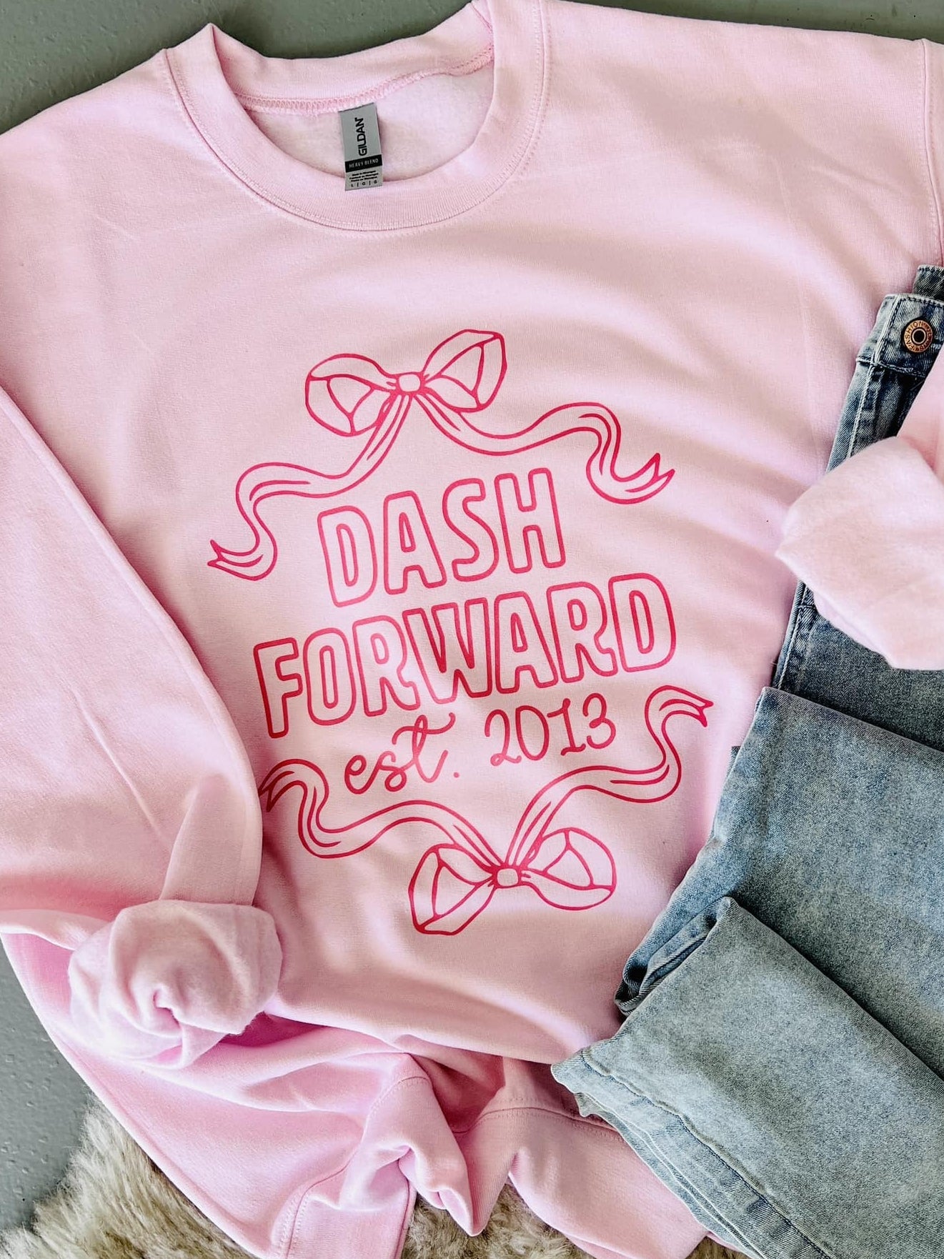 Pink sweatshirt with 'Dash Forward est 2013' text and bow ribbon graphics on a textured surface.
