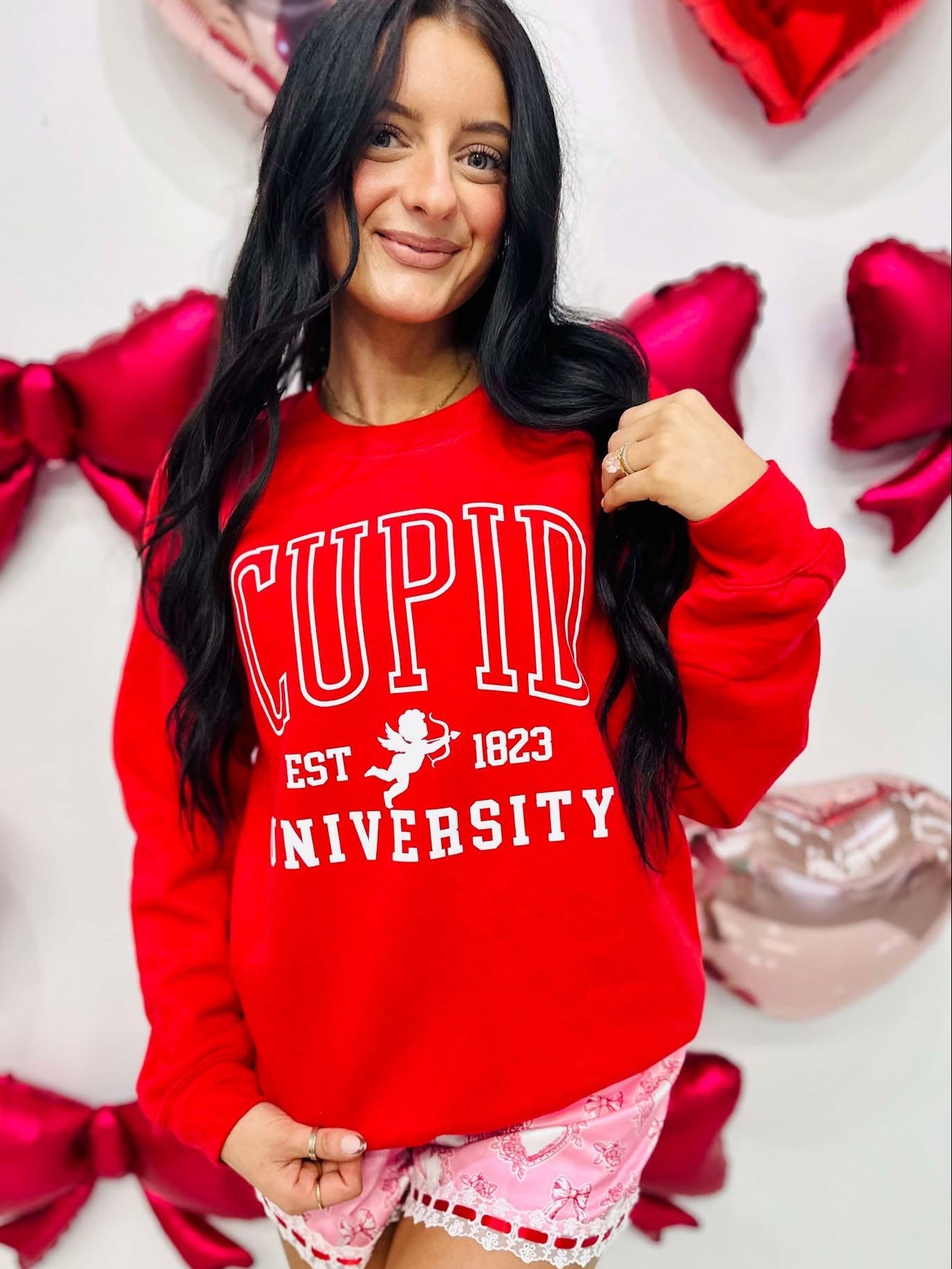 Woman wearing a red 'Cupid University' sweatshirt with heart-shaped decorations in the background.