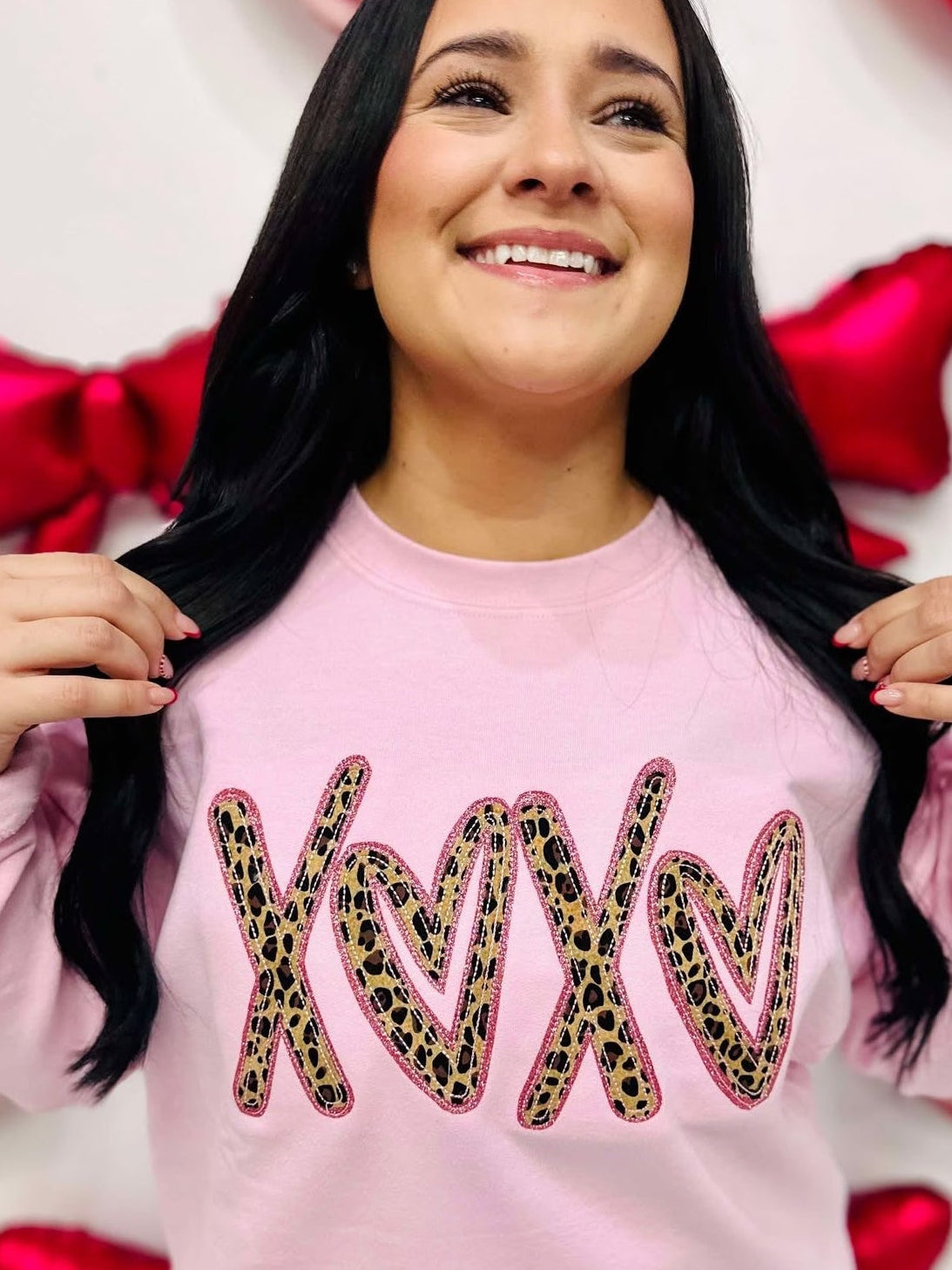 A person wearing a light pink sweatshirt with the word 'XOXO' embroidered in a leopard print style on the front, surrounded by heart-shaped decorations.
