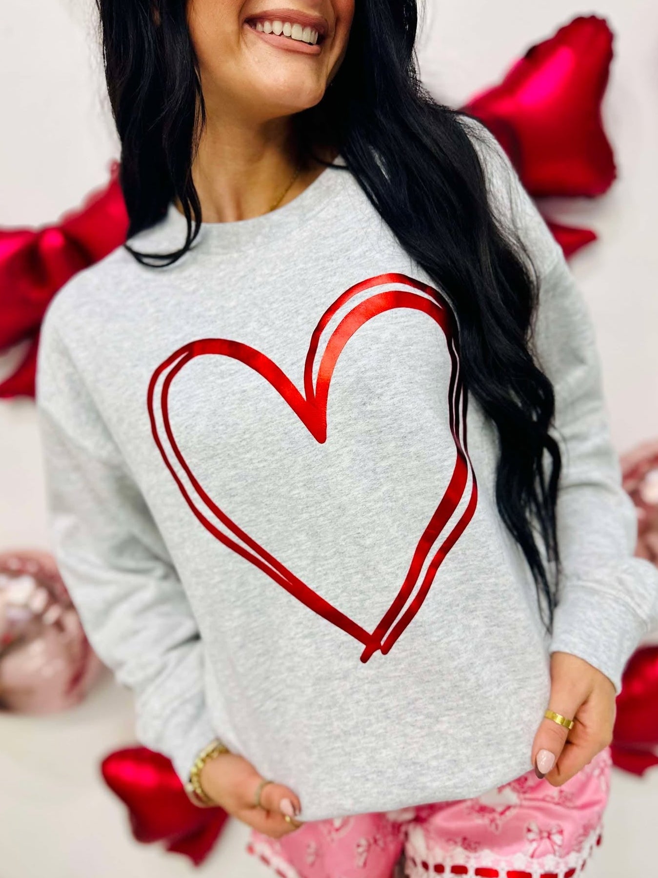 Woman wearing a gray sweatshirt with a red heart design, standing against a white background with red ribbons.