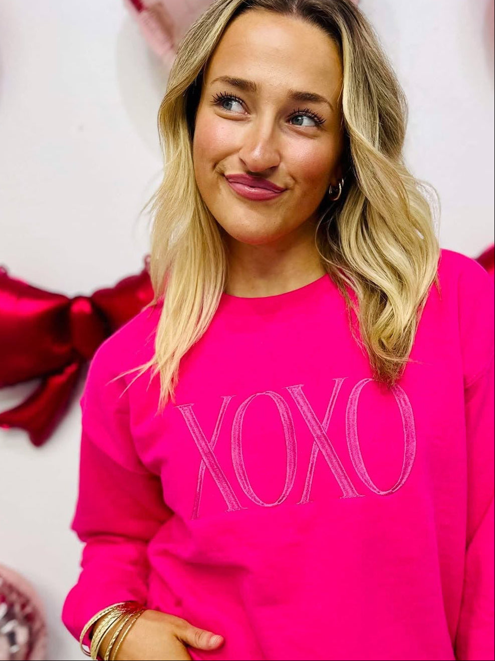 A woman wearing a hot pink embroidered sweatshirt with the text 'XOXO' on it, posing against a background of red and white balloons and decorations.