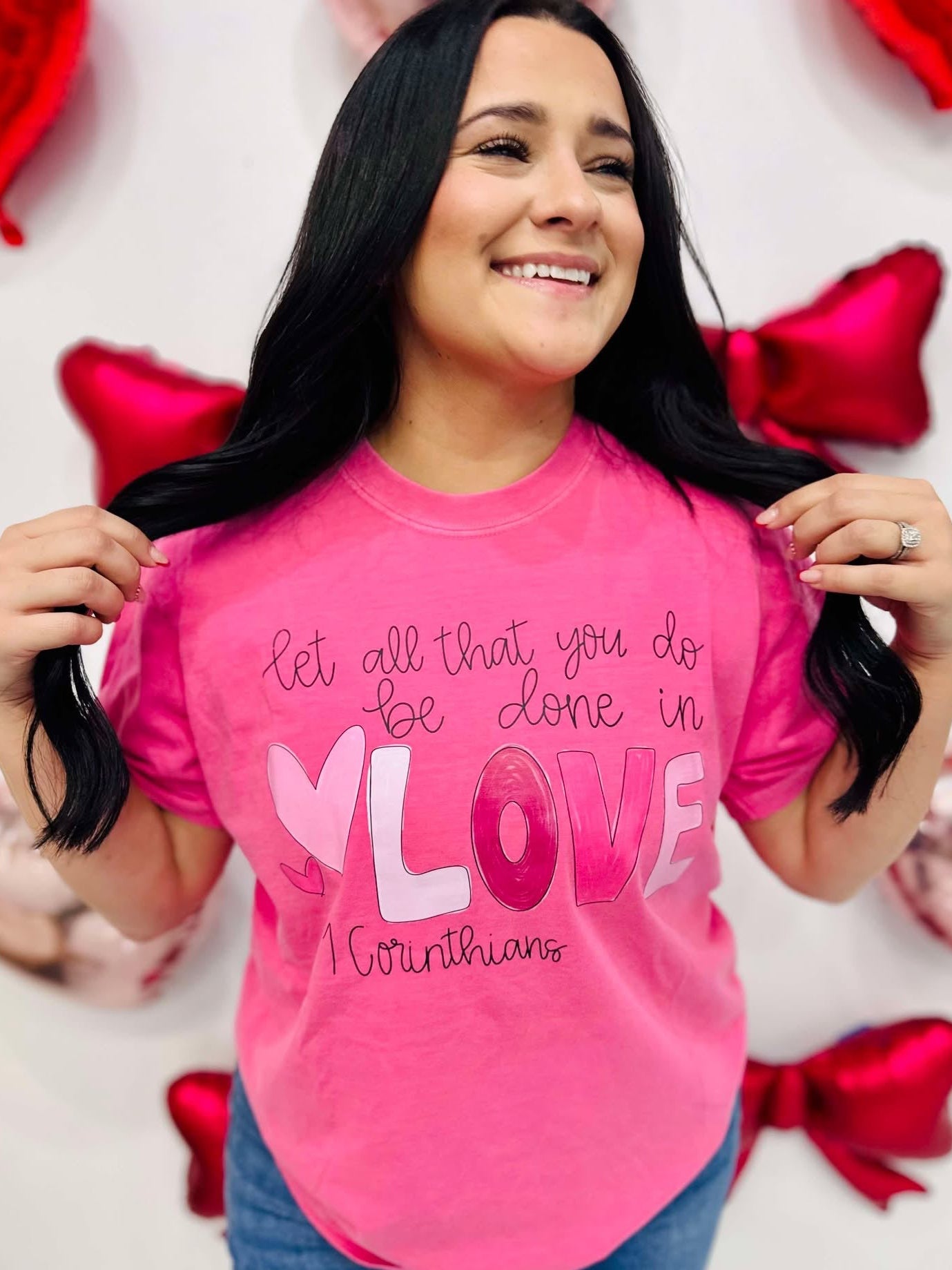 A person wearing a pink t-shirt with the text 'Let all you do be done in LOVE' printed on it, standing in a room with balloons and decorative hearts.