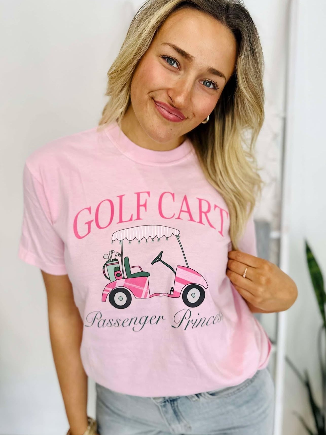A woman wearing a pink t-shirt with 'GOLF CART' and 'PASSENGER PRINCESS' printed on it, standing in a room with a white background.