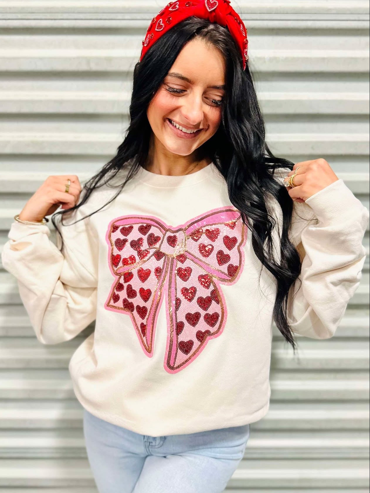 A woman smiling, wearing a beige sweatshirt with a red bow and heart patches, and a red headband.