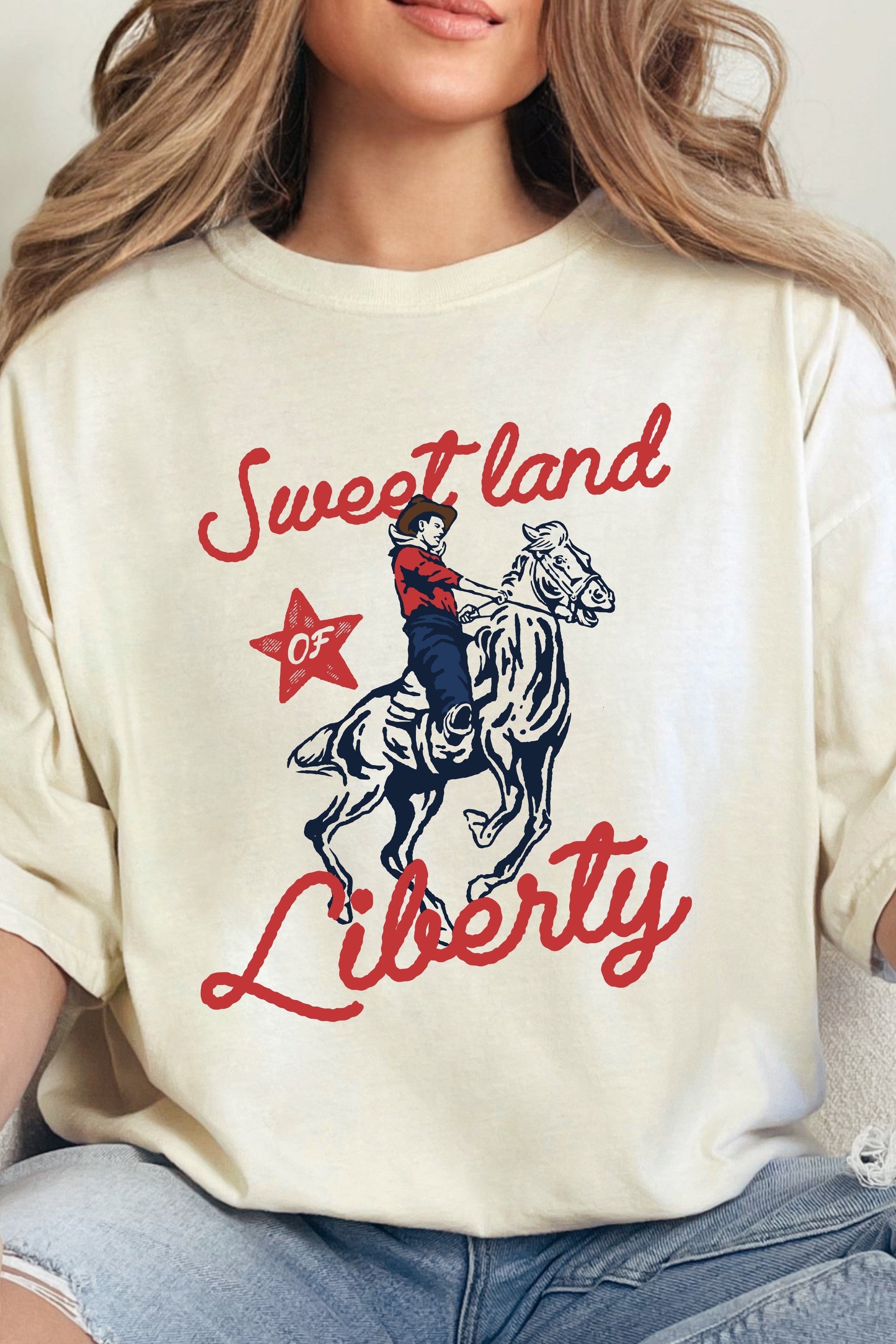 A person wearing an ivory colored graphic tee with the text 'Sweet land Liberty' and a cowboy illustration.
