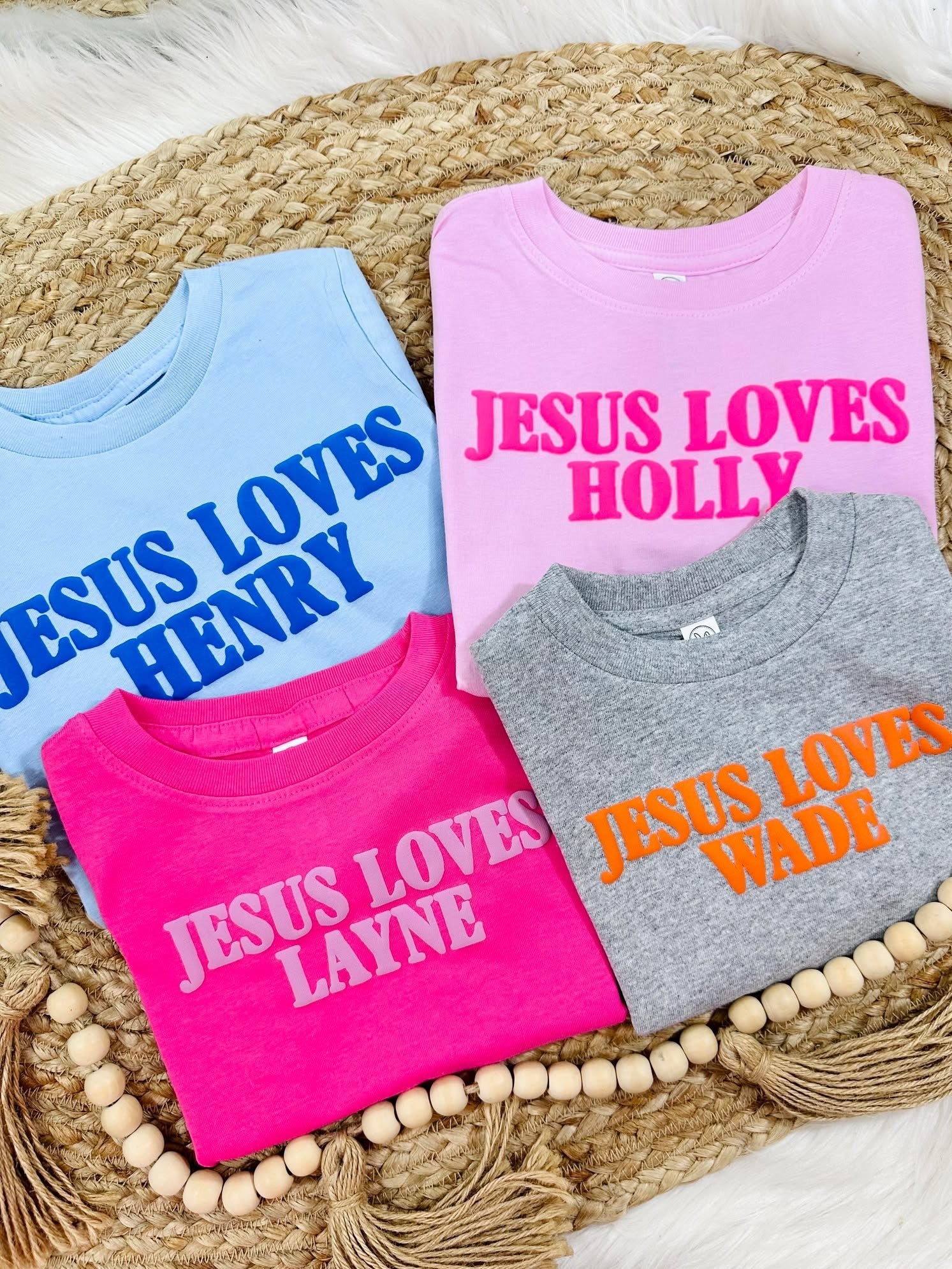 Four children's sweatshirts with Jesus Loves personalized names on a woven mat.