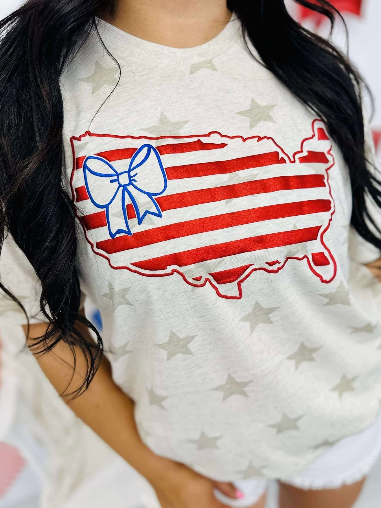 A graphic tee with a patriotic theme featuring an outline of the United States in red and blue colors with a bow on it, against a natural heather star pattern.
