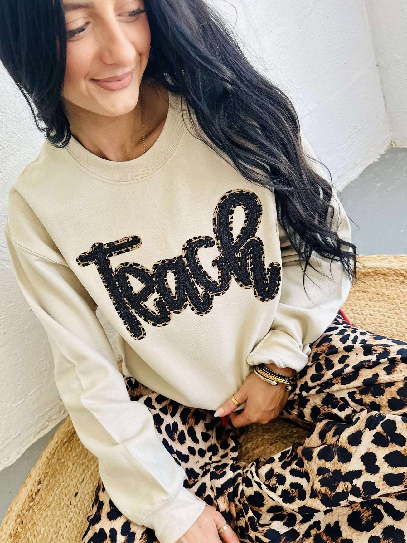 Woman wearing a beige sweatshirt with 'Teach' in black sequins and leopard print pants sitting on a woven chair.