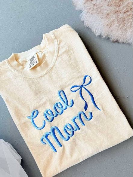 A yellow graphic tee shirt with blue embroidered lettering that reads 'Cool Mom' with a bow detail above the text.