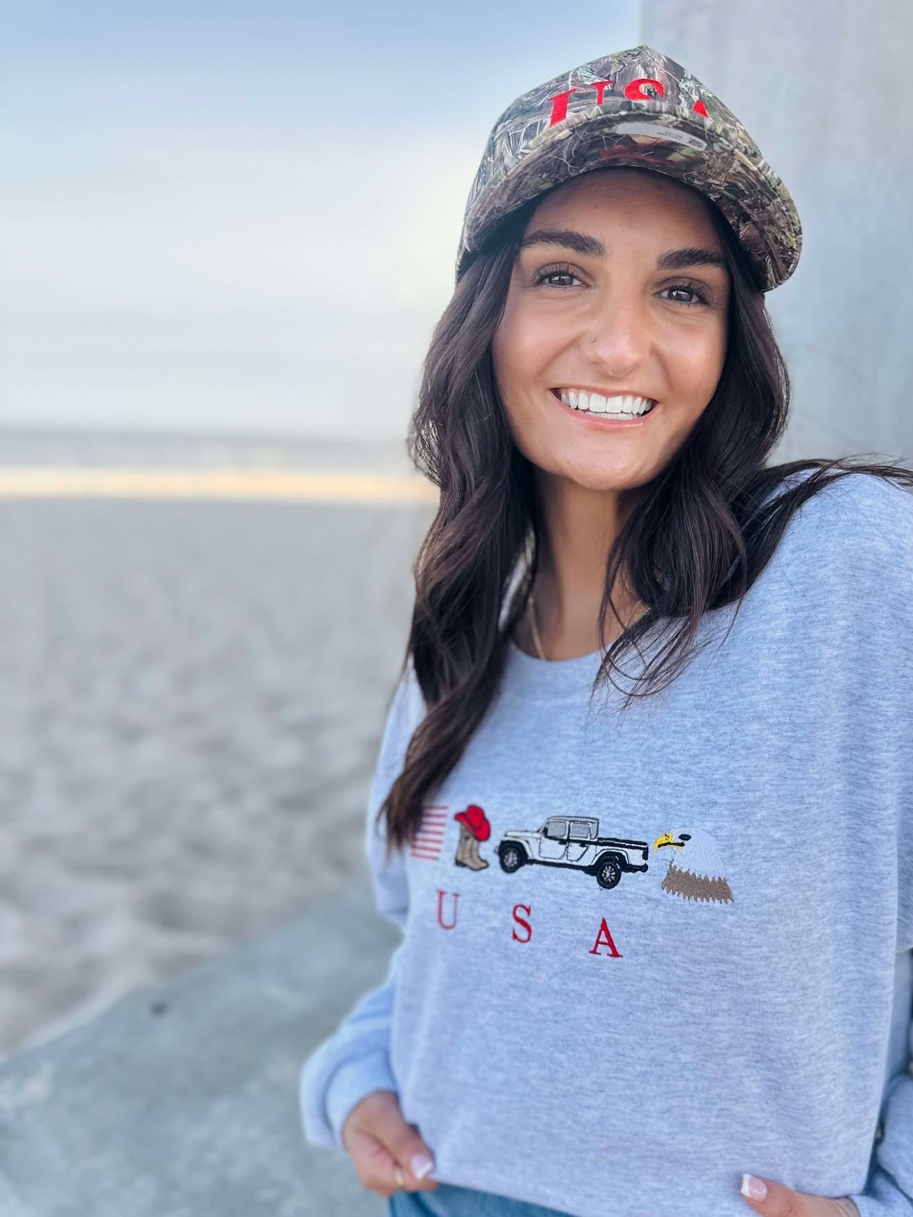 Woman wearing a light blue sweatshirt with an eagle, cowboy boots, truck and usa flag graphic design, sitting on a beach.