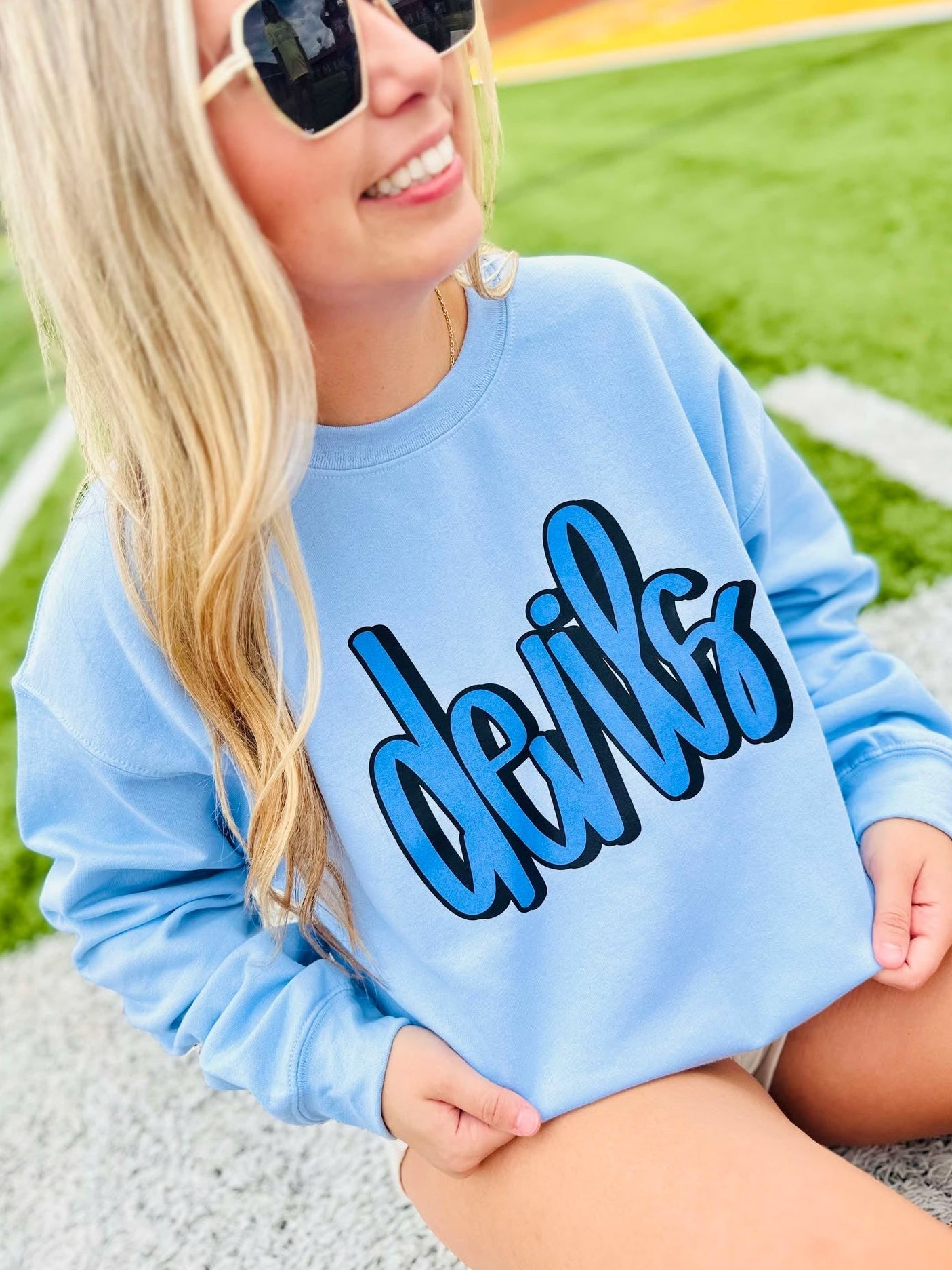 Person wearing a light blue sweatshirt with 'devils' printed on it, sitting on a grassy field.
