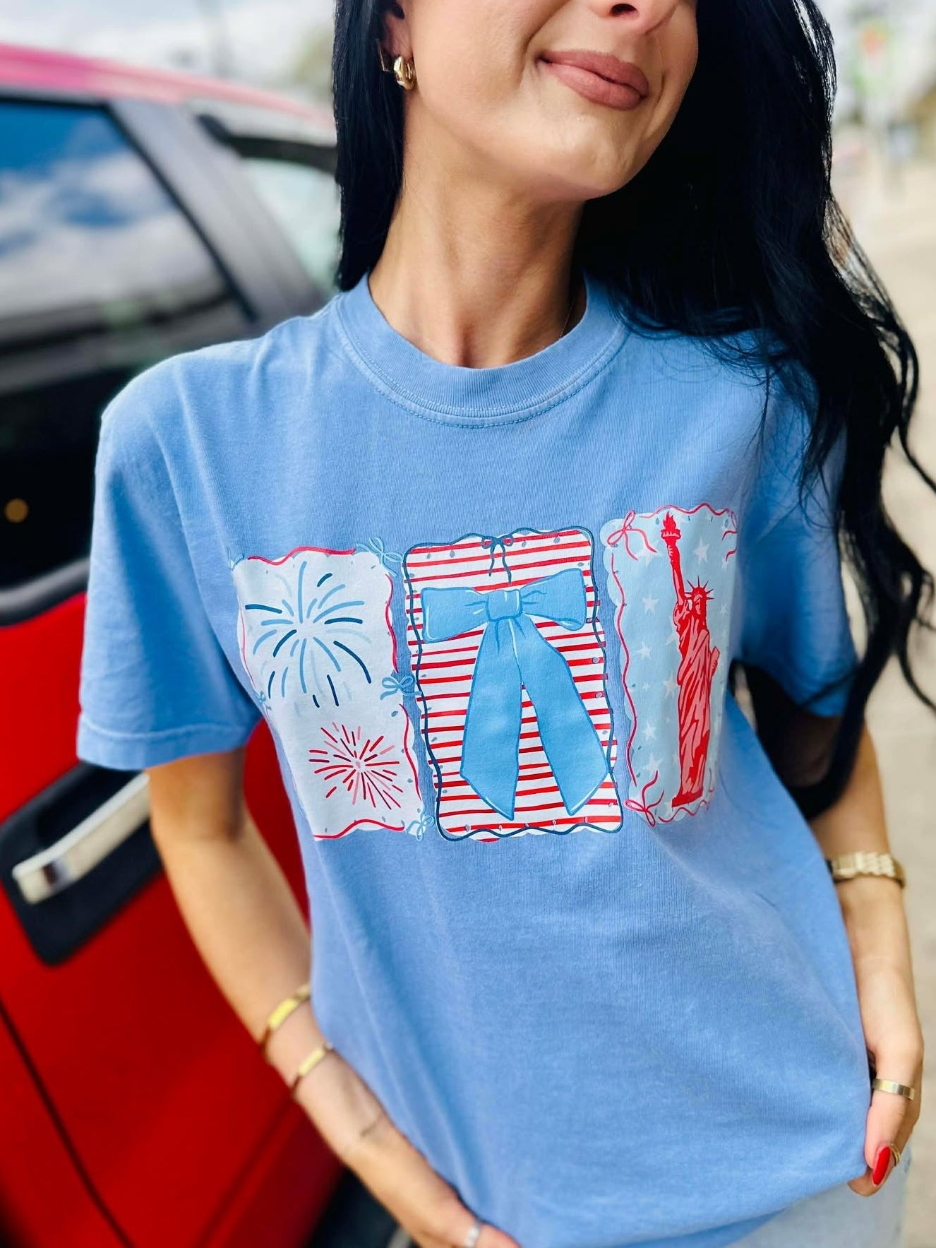 Woman wearing a blue t-shirt with graphic designs, standing next to a red car.