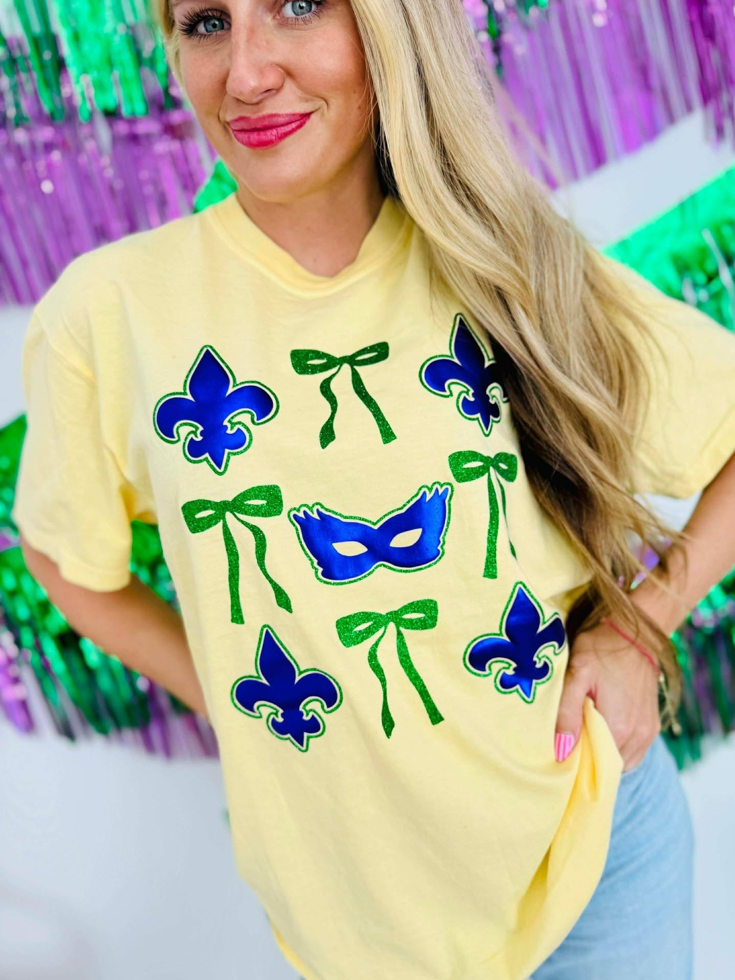 Woman wearing a yellow t-shirt with purple masks and green ribbons in front of a colorful background for Mardi Gras