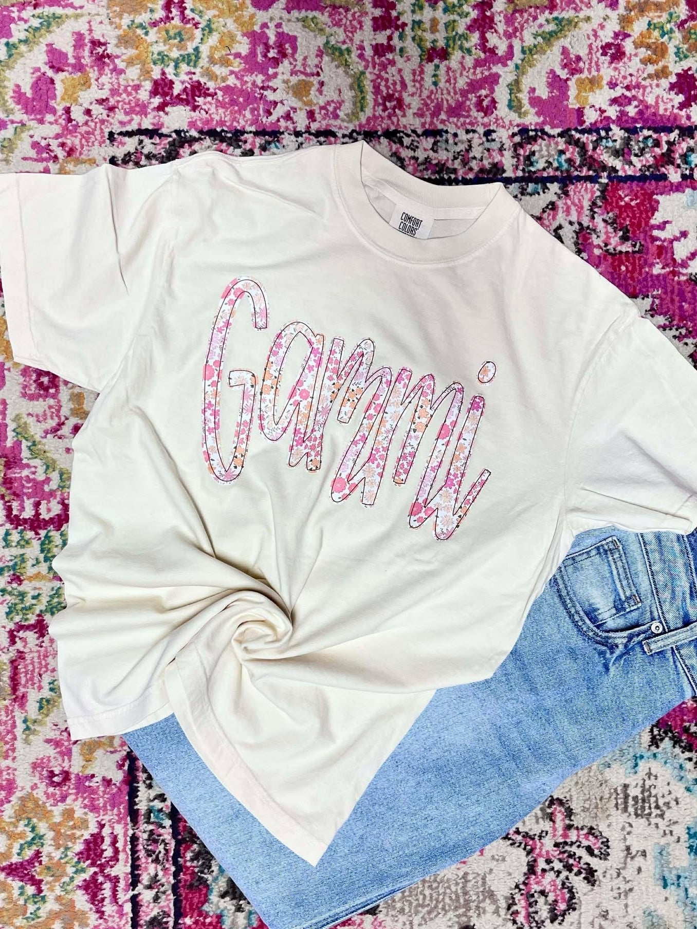 White t-shirt with colorful 'Gammi' text on a multicolored patterned surface
