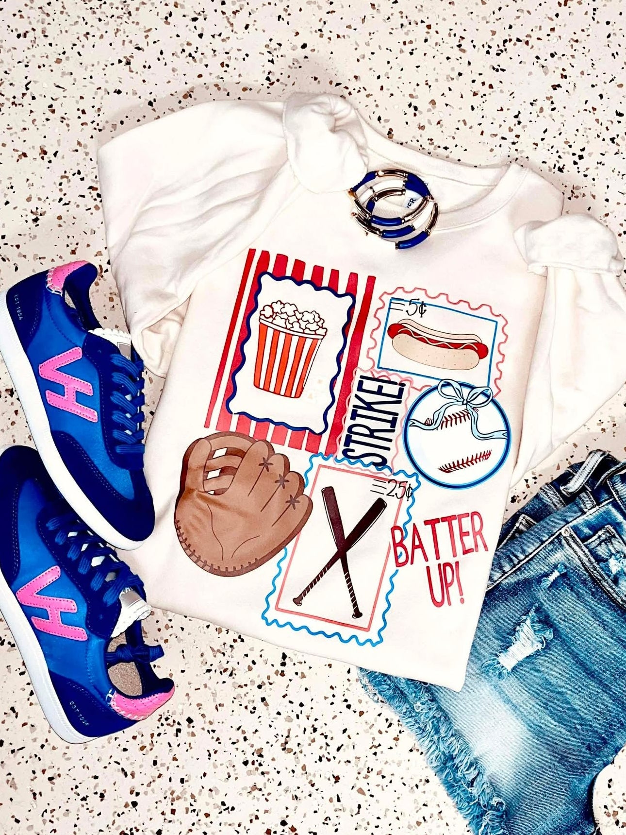 White t-shirt with sports-themed design, blue sneakers, baseball glove, and denim shorts on a speckled surface.