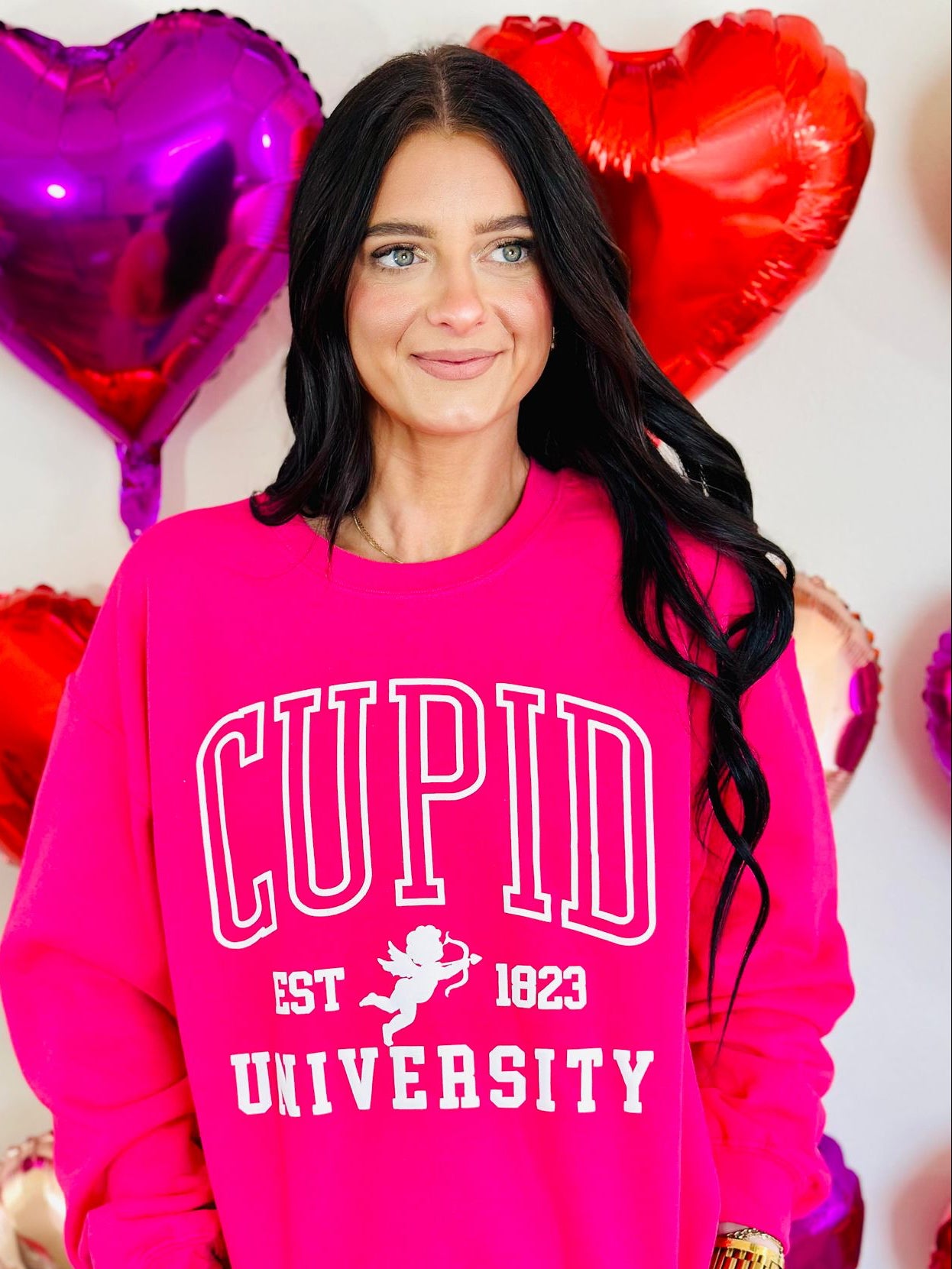 A person wearing a pink sweatshirt with the text 'CUPID UNSUCCESSFUL SINCE 1823' and a graphic of a cupid, surrounded by heart-shaped balloons.