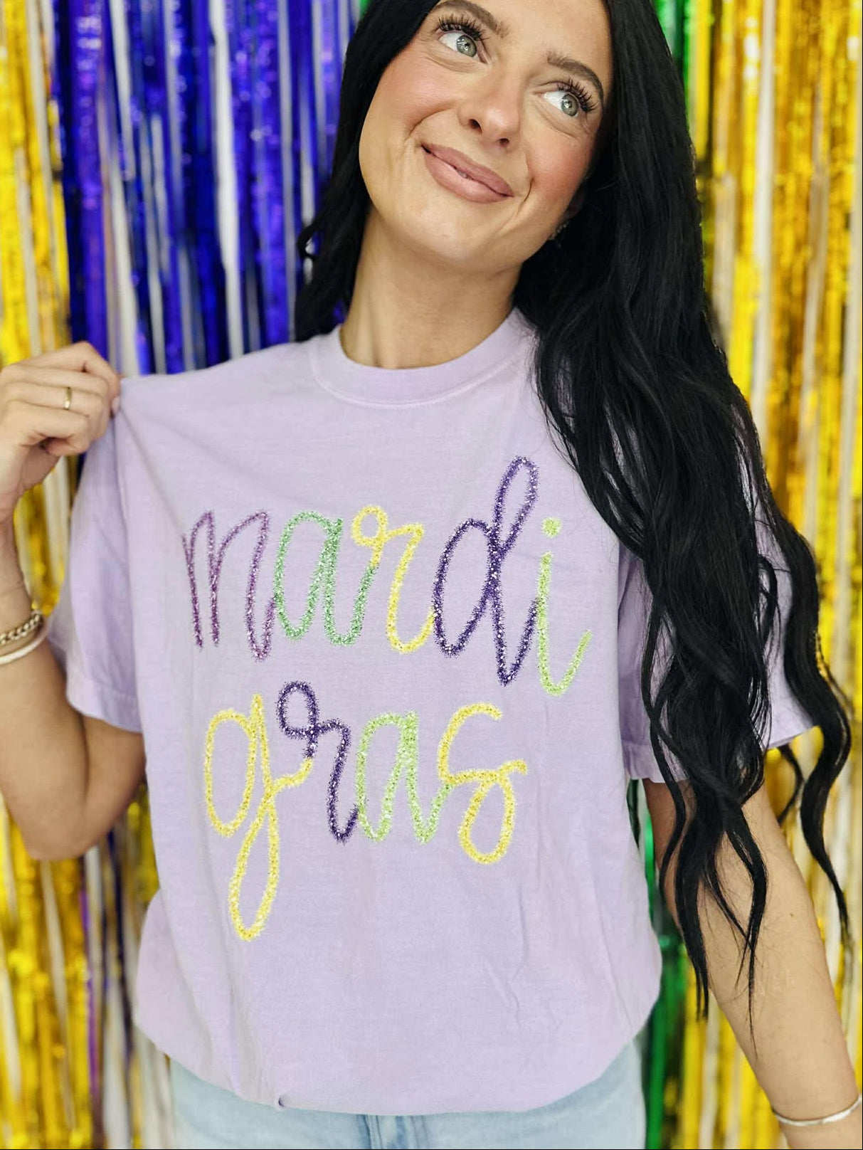 A person holding up a purple t-shirt with 'mardi gras' printed in yellow and neon green tinsel text.