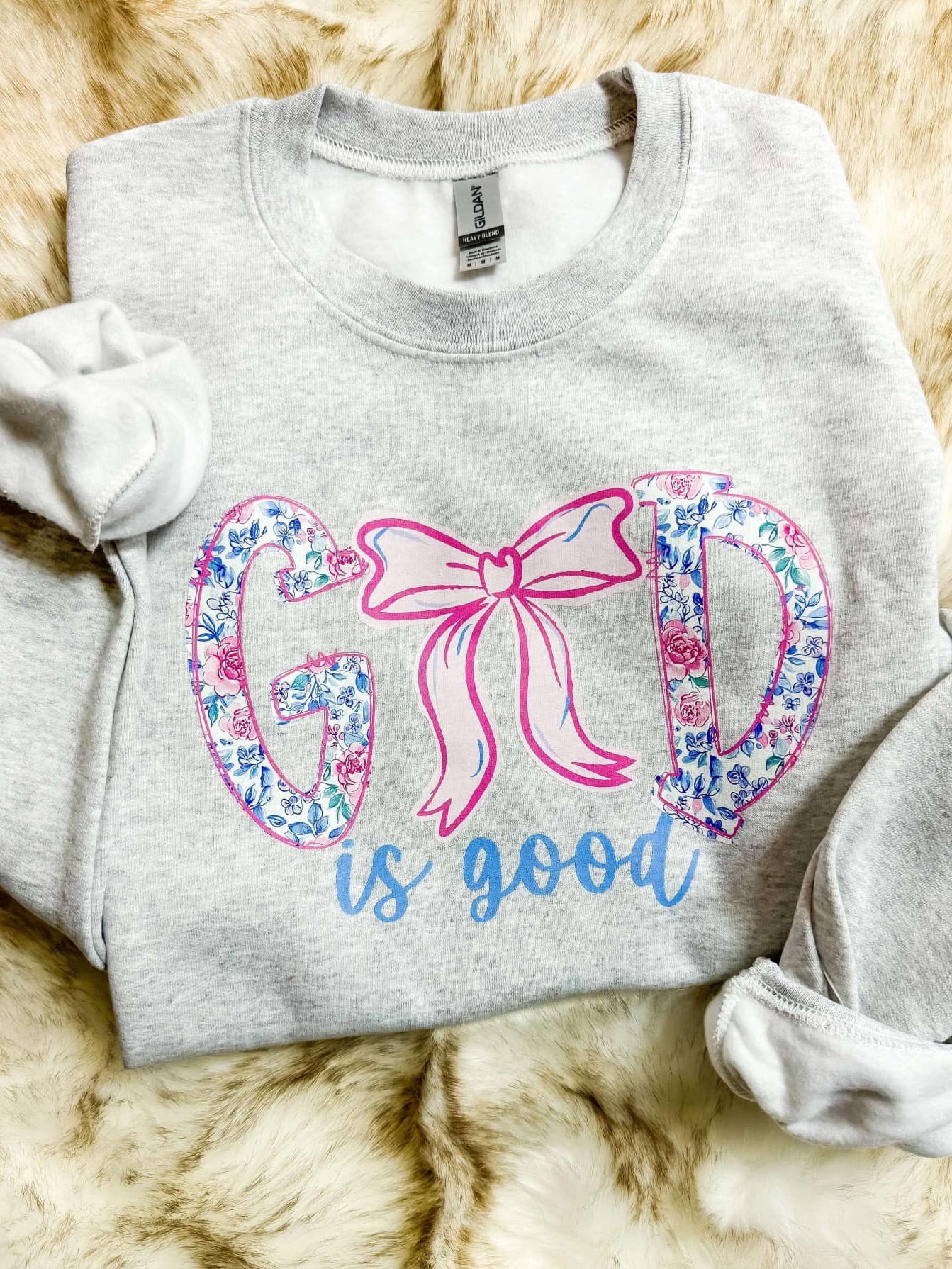 Ash grey sweatshirt with the phrase 'God is good' printed in a floral ribbon style in pink and blue.