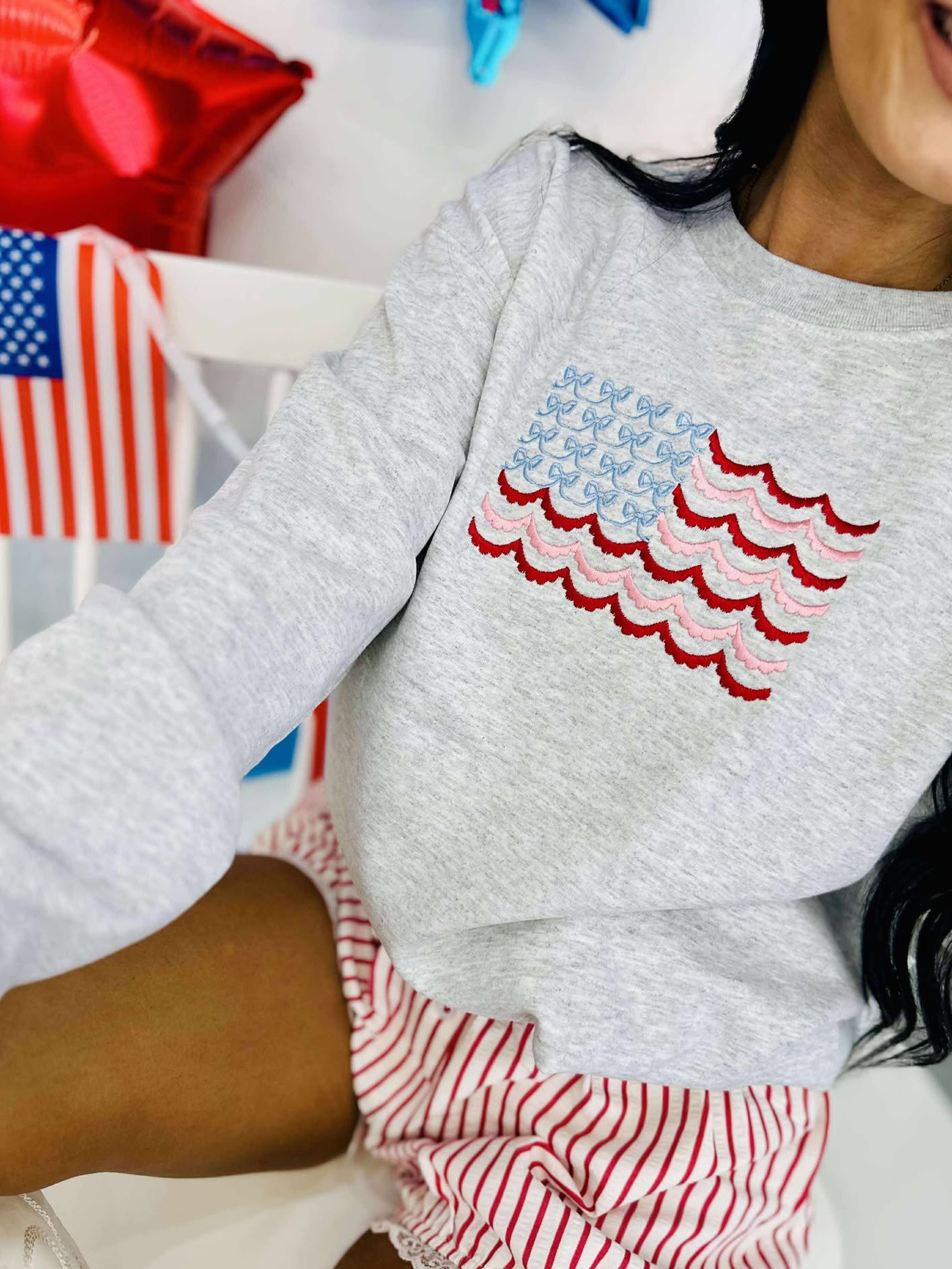 A sweatshirt with an embroidered American flag design in red and blue on a gray background, worn by a person seated in front of a small table decorated with American flags and related merchandise.
