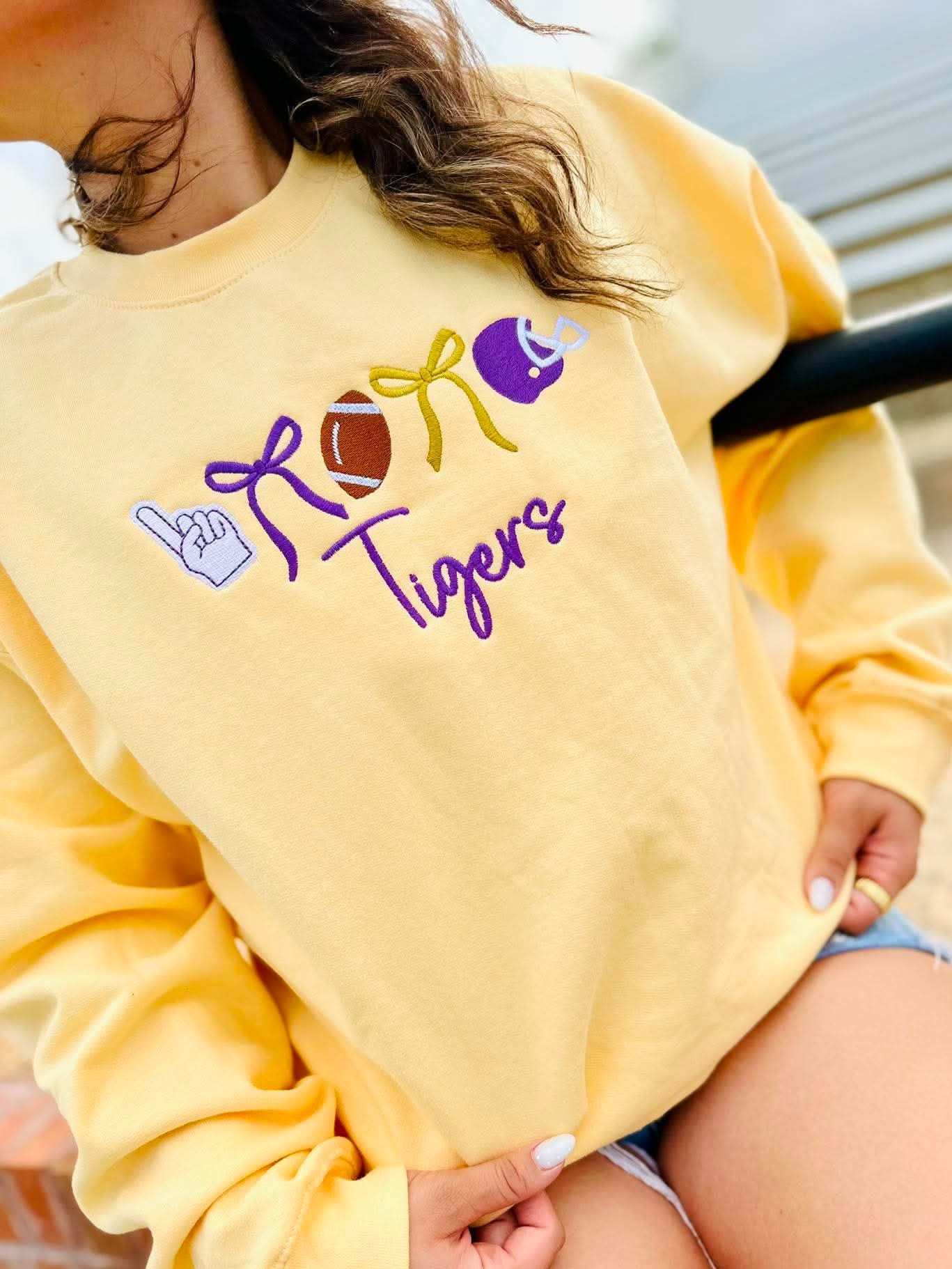 A person wearing a pale yellow sweatshirt with a 'ROAR Tigers' graphic featuring purple, yellow, and brown football-themed illustrations, paired with denim shorts.