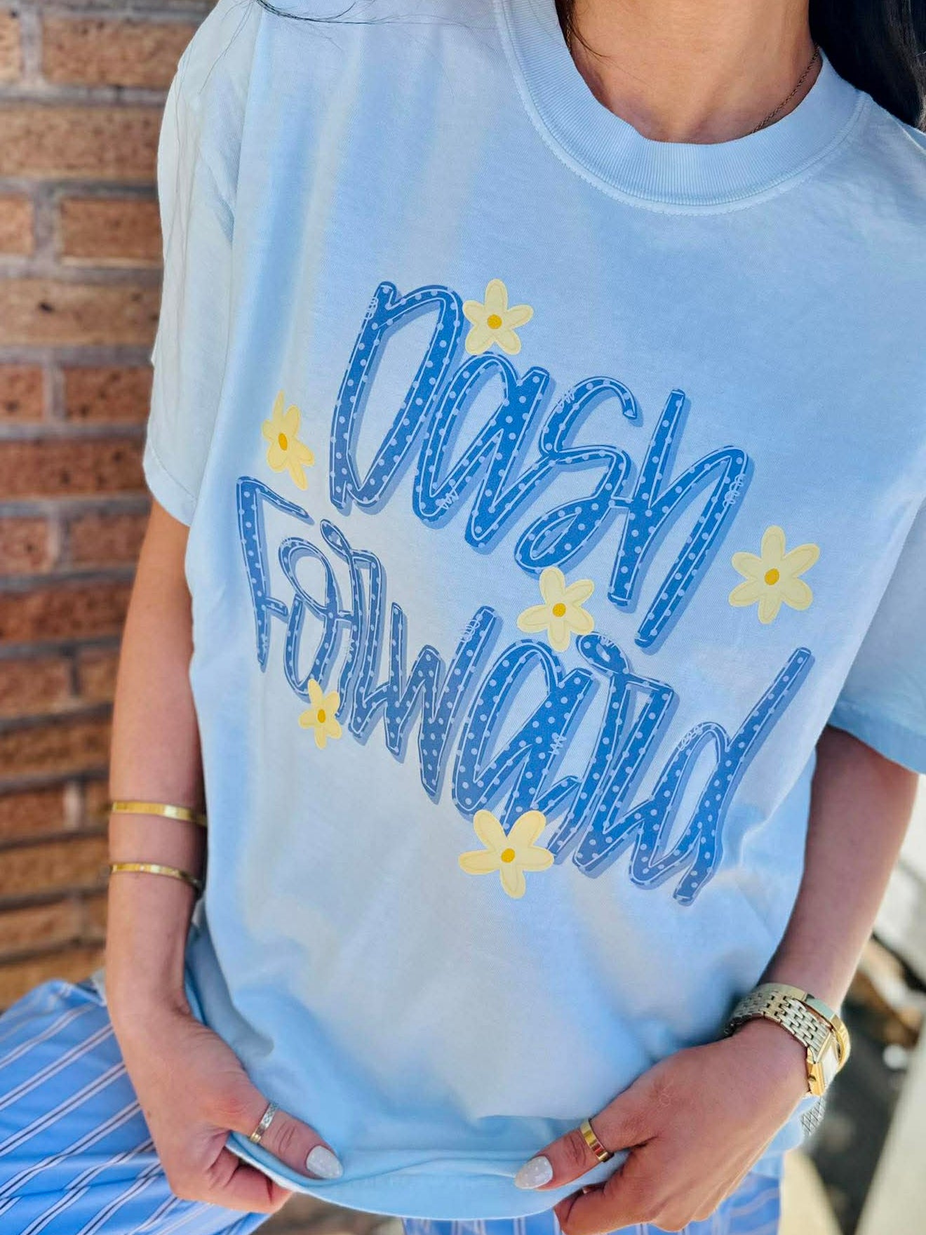 Person wearing a light blue t-shirt with 'Dash Forward' text and stars, standing against a brick wall.