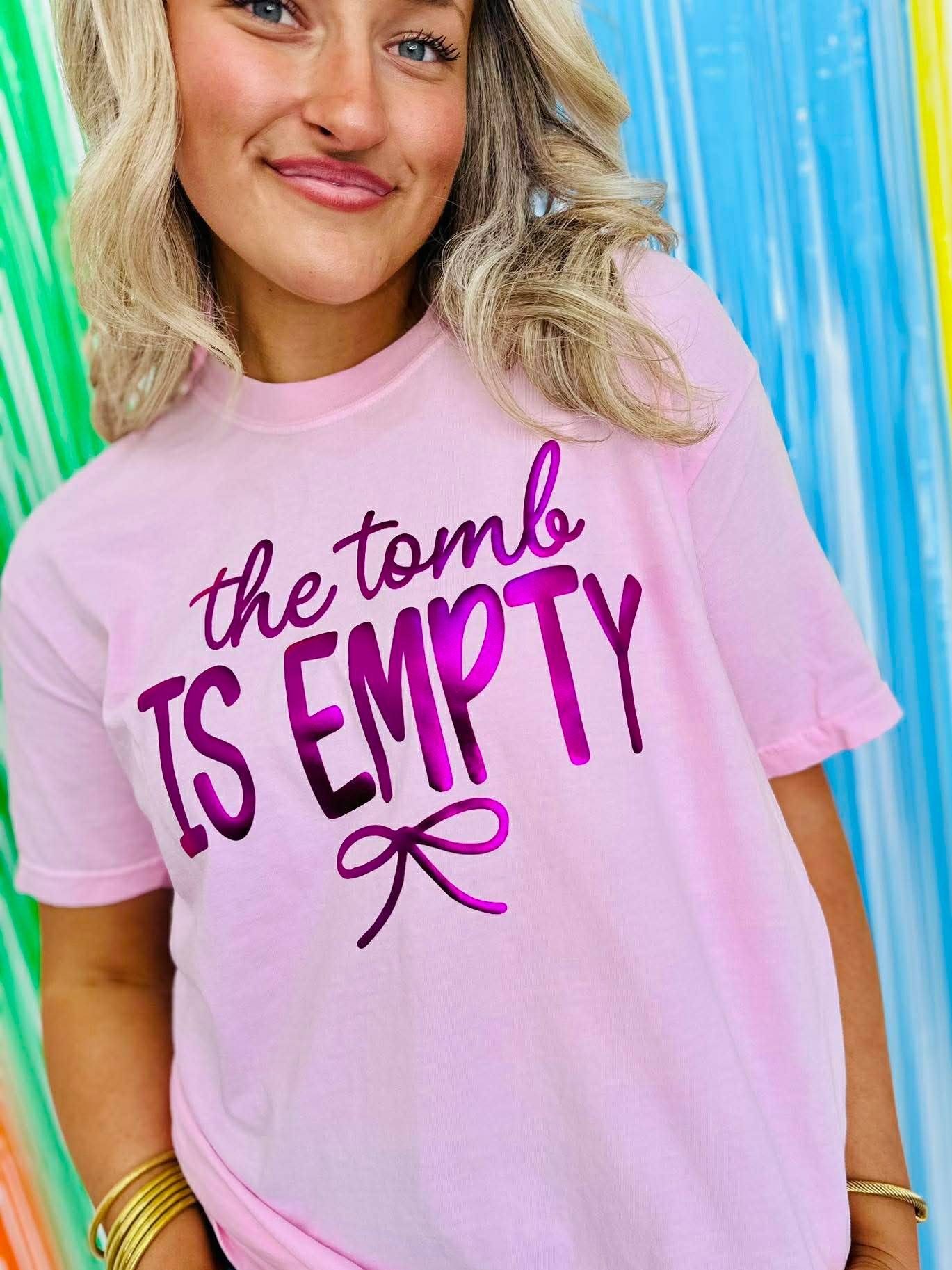 Person wearing a pink t-shirt with 'the tomb is empty' puff metallic text in front of a colorful striped background