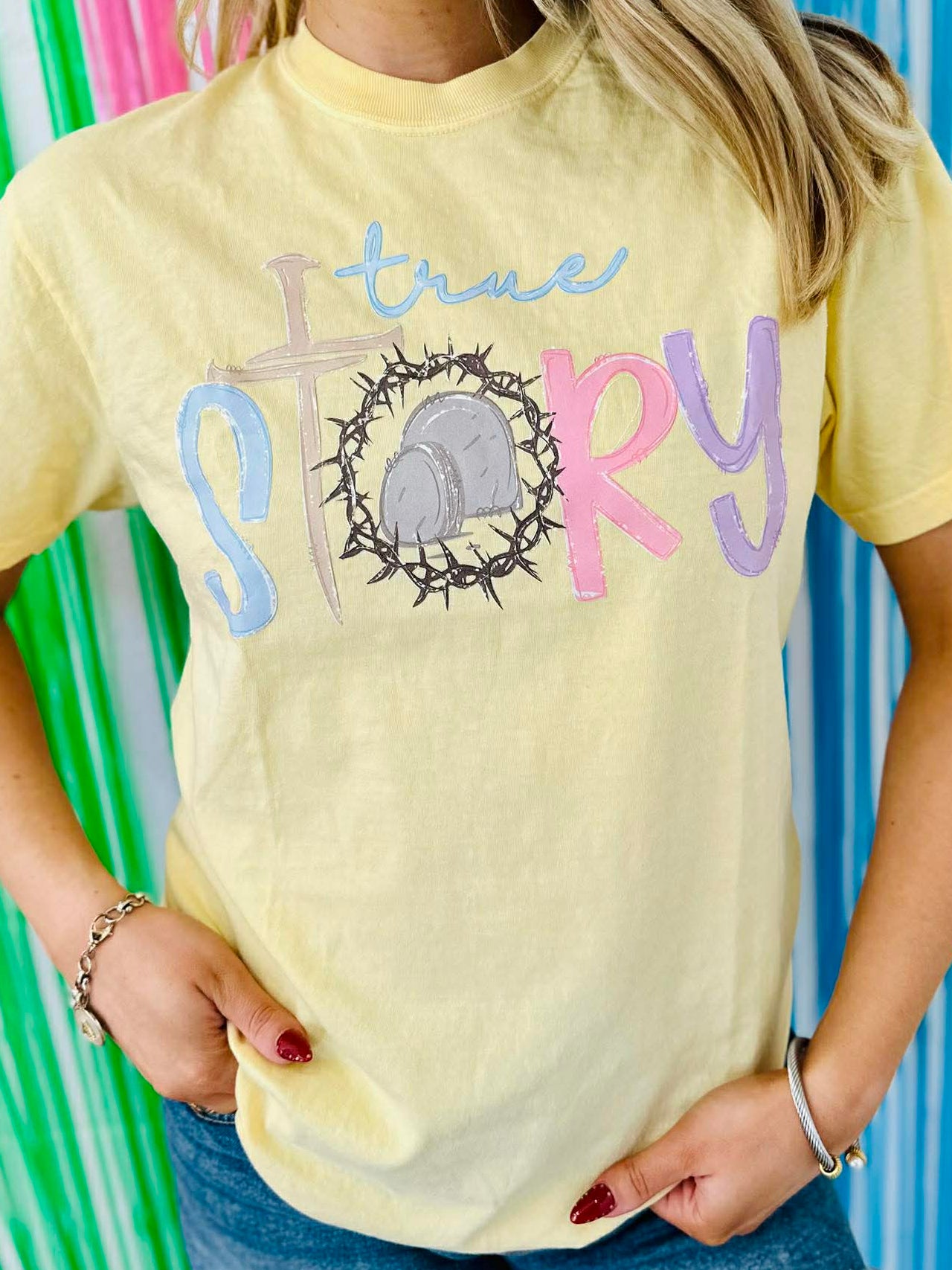 Person wearing a yellow t-shirt with 'true story' text with crown of thorns and tomb against a colorful striped background