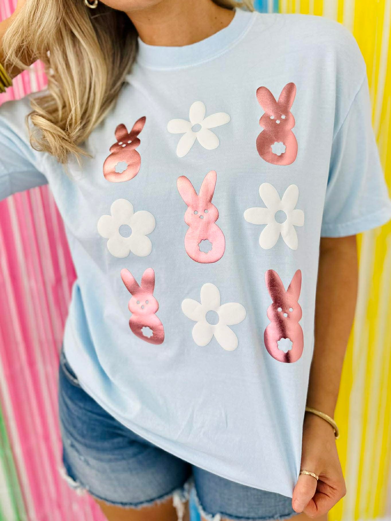 Person wearing a light blue t-shirt with rose gold peep bunny and white puff vinyl flower designs against a colorful striped background.